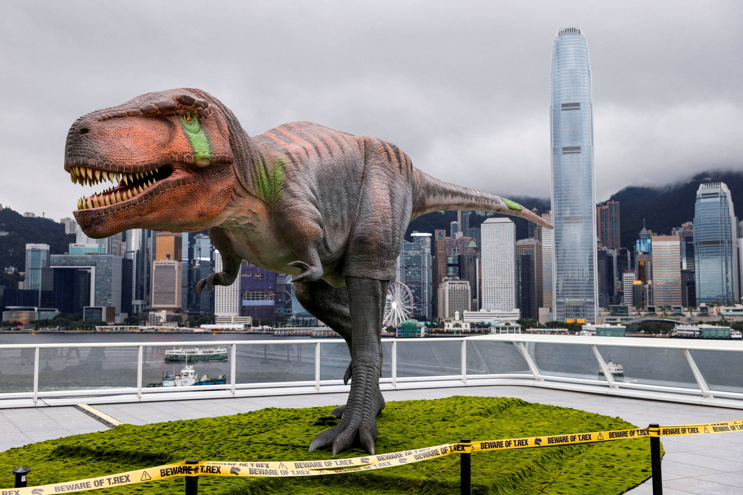 Dinossauro animatrônico de grande porte em plataforma com grama artificial, cercado por fita amarela de advertência, com arranha-céus e céu nublado ao fundo.