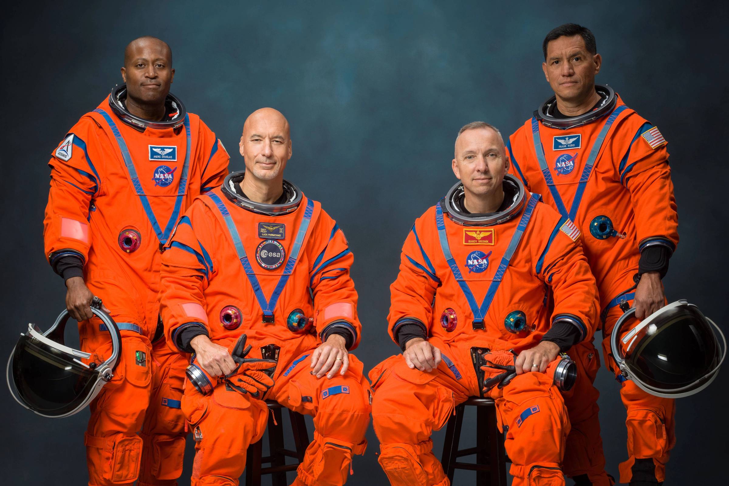 Quatro astronautas posam em trajes espaciais laranja com detalhes azuis e emblemas da NASA. Dois estão sentados em bancos, segurando capacetes pretos, e dois estão em pé atrás deles, contra fundo neutro escuro.