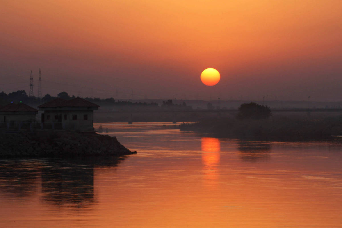 Sol alaranjado próximo ao horizonte sobre rio calmo, refletindo luz dourada na água. Margens com vegetação e construções escuras ao fundo.
