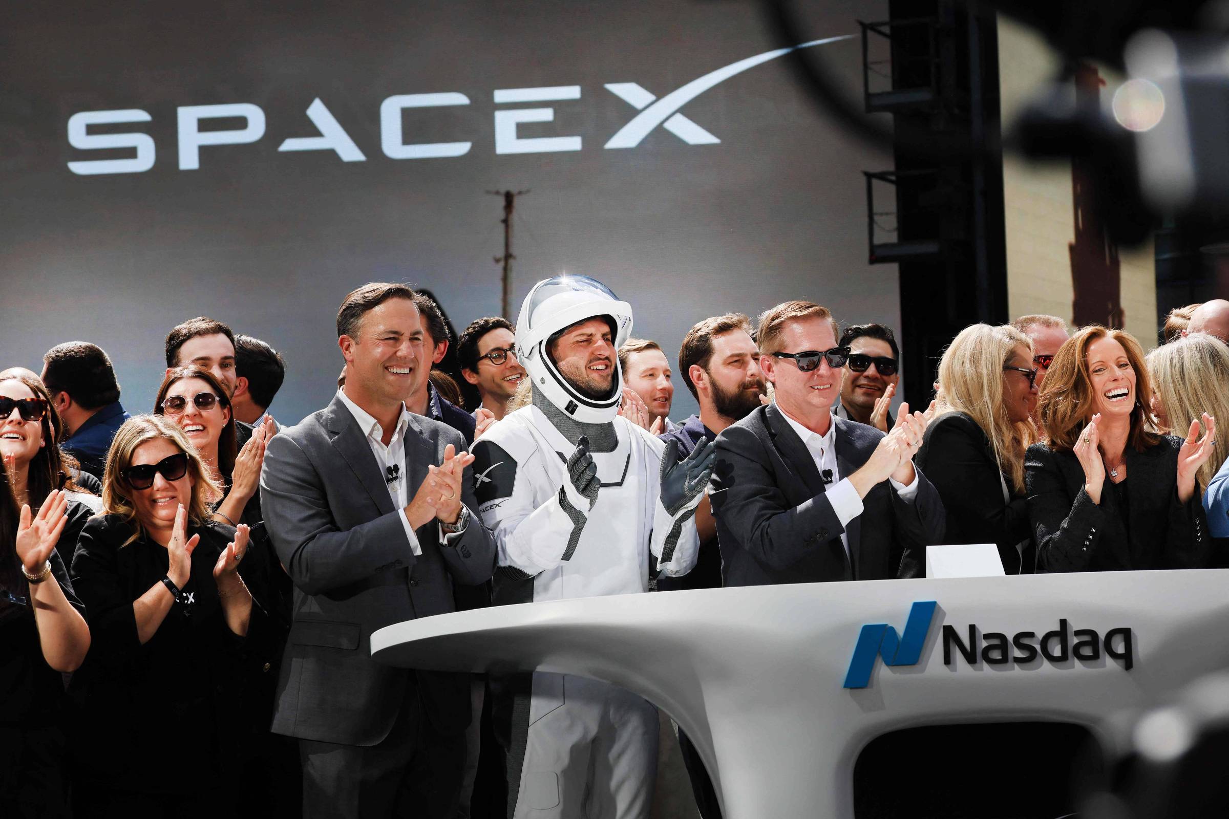 Astronauta em traje espacial da SpaceX e grupo de pessoas aplaudem em evento da Nasdaq. Logo da SpaceX iluminado ao fundo.