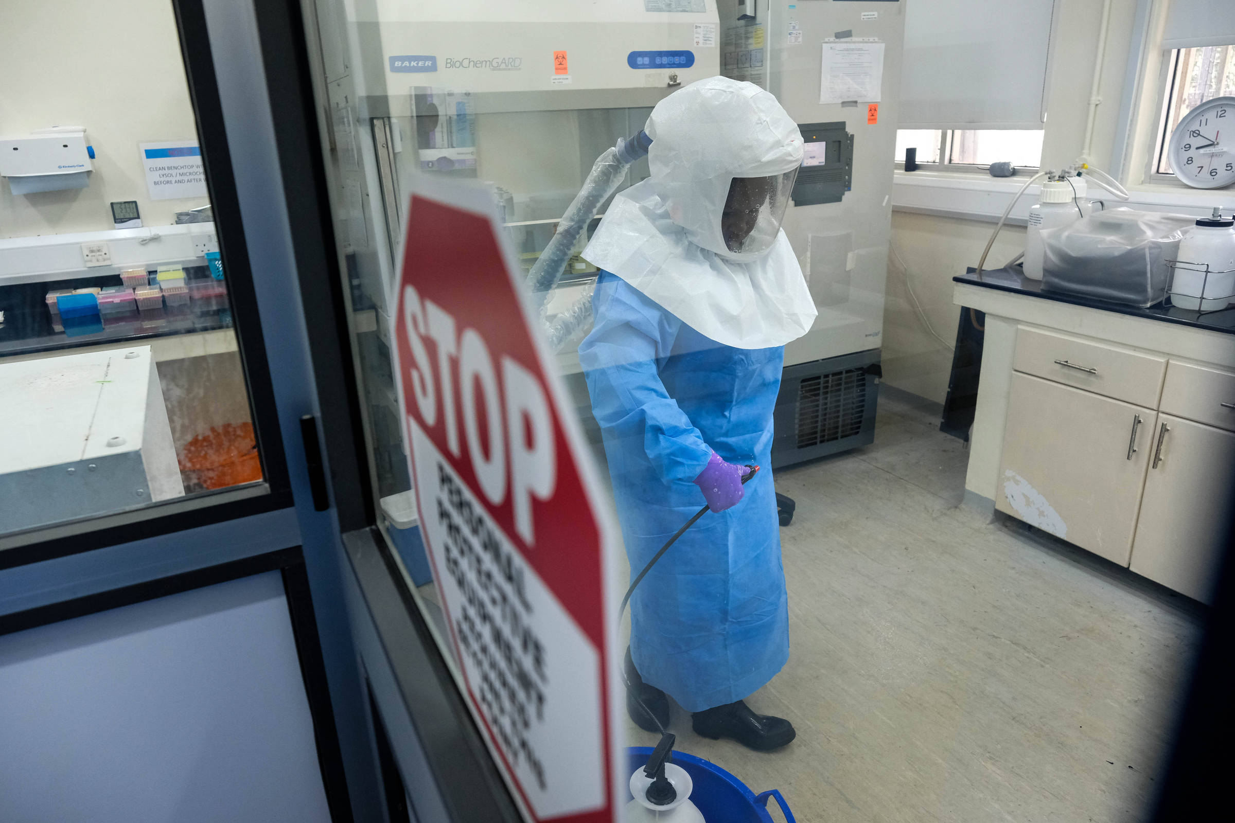 Pesquisador usando traje de proteção completo azul e máscara com capuz transparente dentro de laboratório. Ambiente contém equipamentos científicos e sinal de aviso