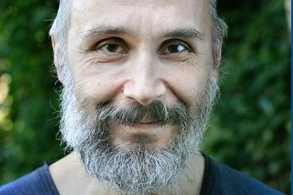 Homem de meia-idade com barba e cabelo grisalho, usando camiseta escura, posando em ambiente externo com vegetação ao fundo.