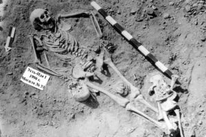 Esqueleto humano completo em posição estendida em solo de escavação arqueológica, com régua de medição ao lado e placa identificadora com texto em cirílico.