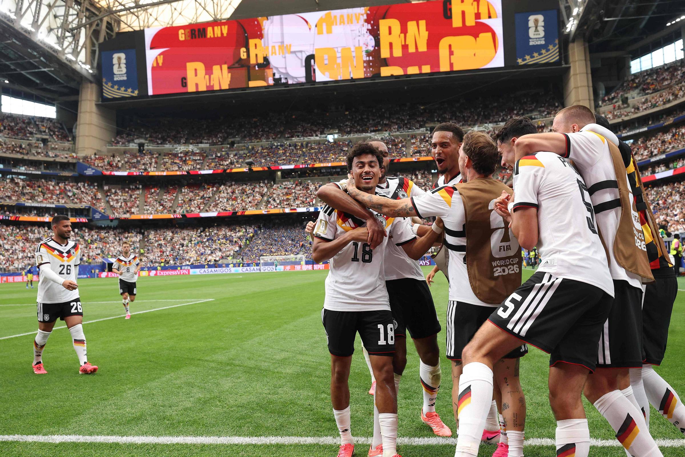 Jogadores da seleção alemã de futebol comemoram gol no campo durante partida em estádio lotado. Um grupo abraça o autor do gol, enquanto outros jogadores se aproximam para celebrar.