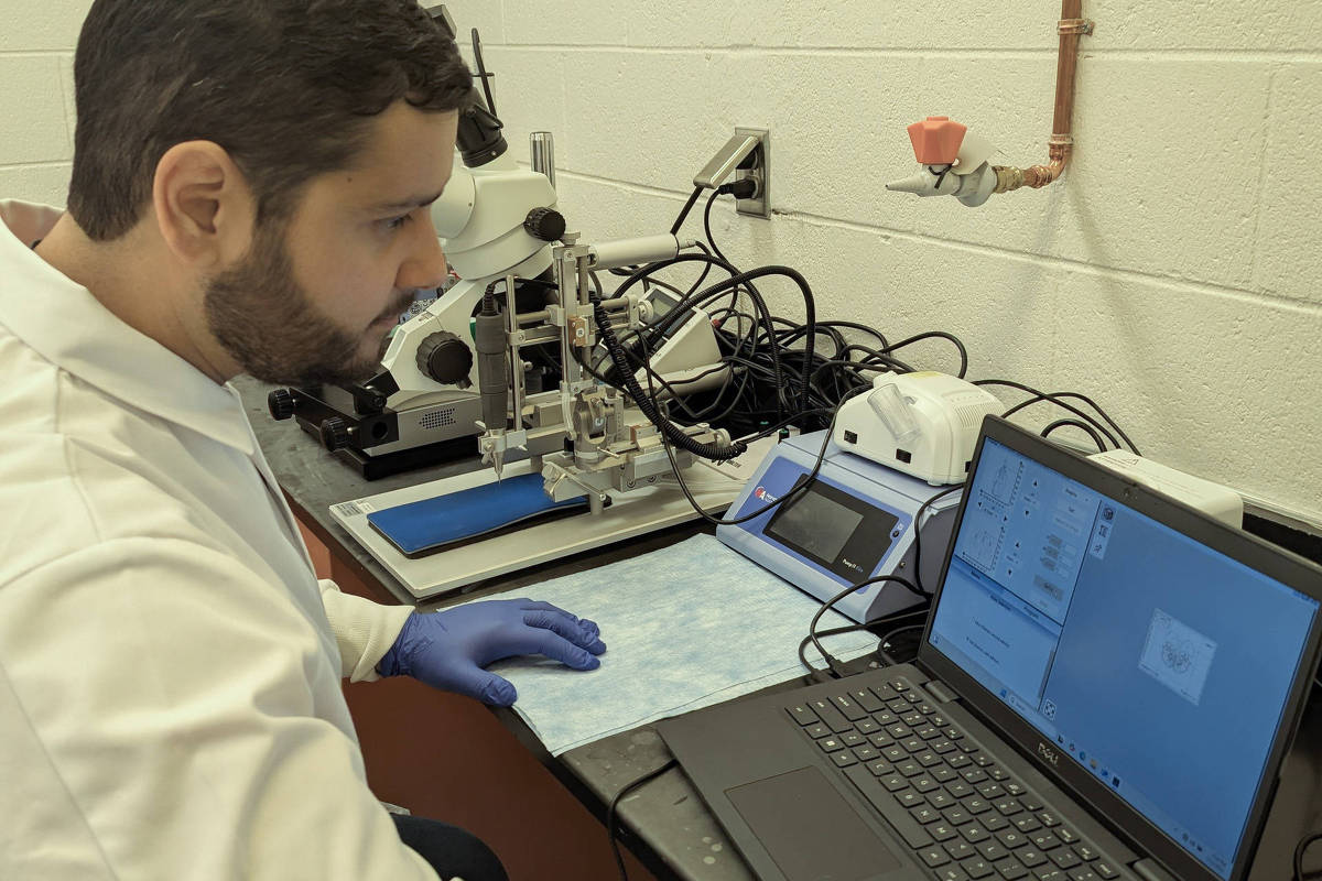Homem com jaleco branco e luvas azuis sentado em bancada de laboratório, operando laptop conectado a equipamento científico com múltiplos cabos e microscópio ao fundo.