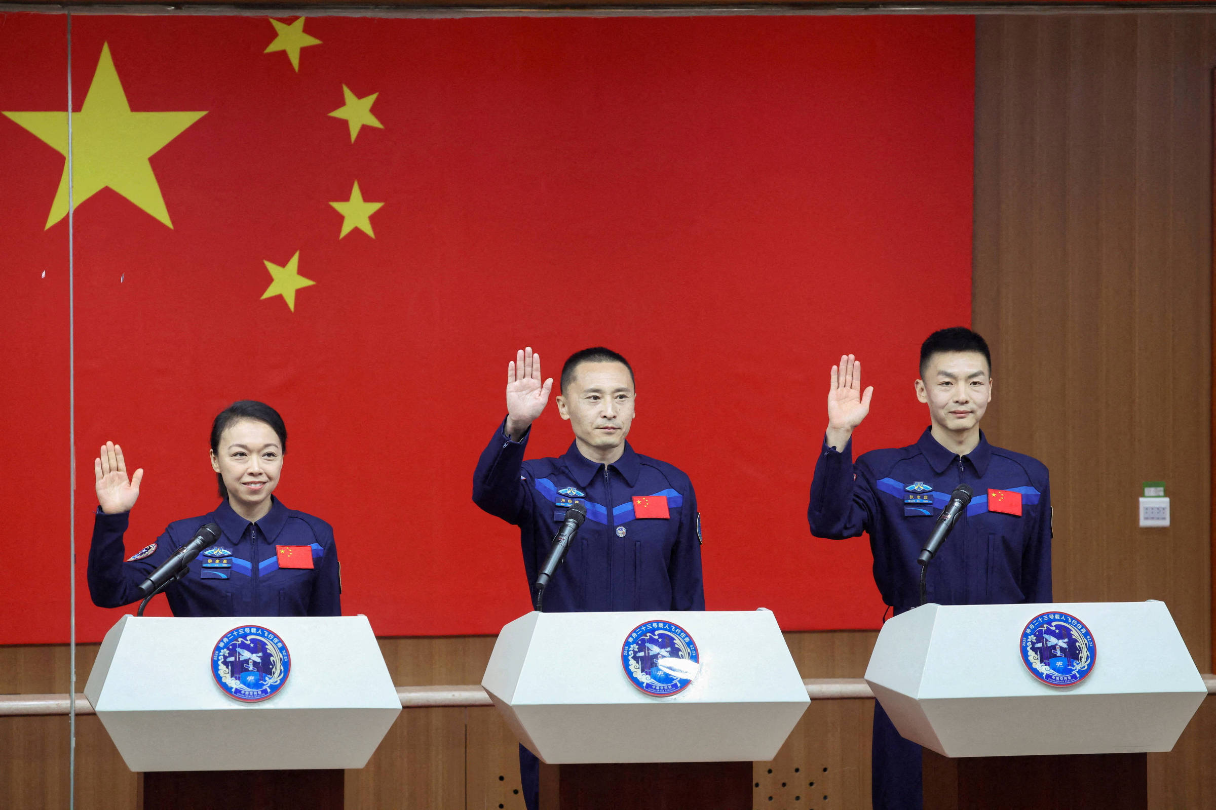 Três astronautas chineses em uniformes azuis com bandeira da China no peito levantam a mão direita em gesto de juramento diante de microfones. Ao fundo, grande bandeira da China vermelha com estrelas amarelas.