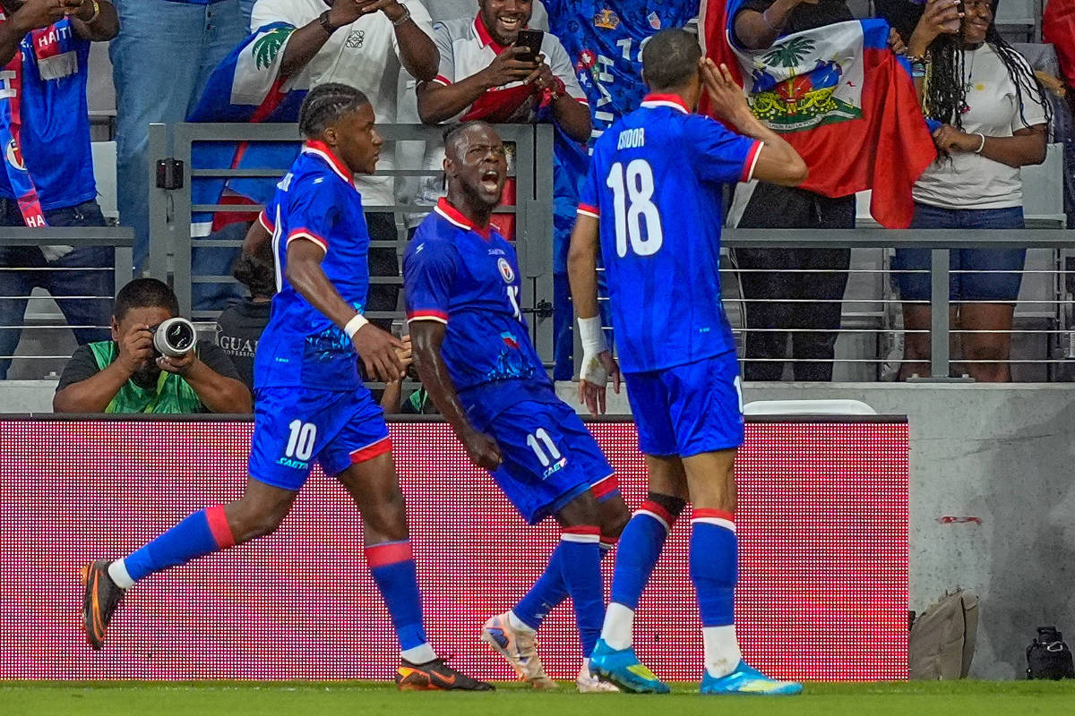 Três jogadores da seleção haitiana de futebol comemoram gol no campo enquanto torcedores vibram nas arquibancadas com bandeiras do Haiti.