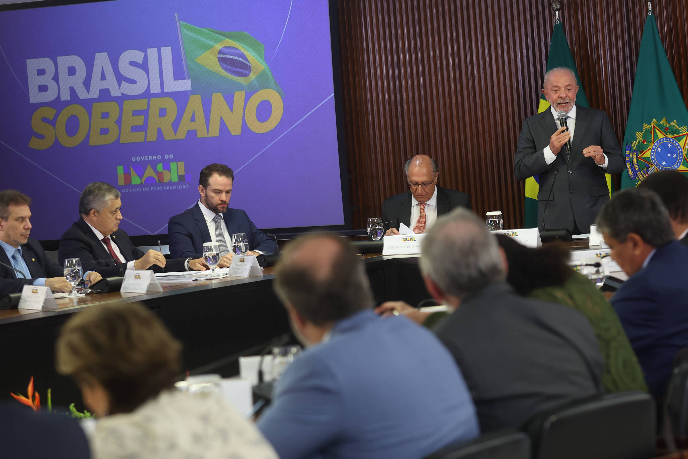 Homem em pé fala ao microfone em reunião formal com várias pessoas sentadas ao redor de mesa. Ao fundo, painel com texto