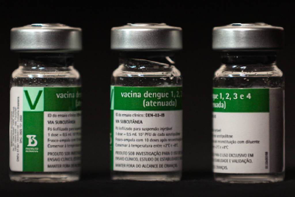 Três frascos de vidro com tampas metálicas contendo vacina contra dengue, alinhados lado a lado sobre superfície preta. Rótulos verdes e brancos indicam informações do medicamento.