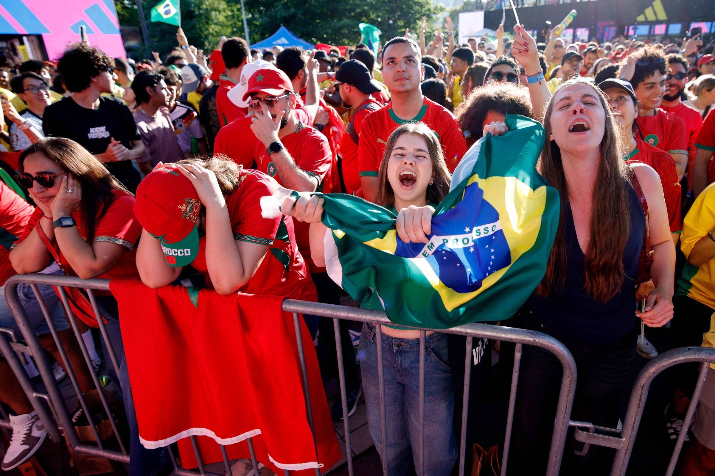 Grupo de torcedores brasileiros atrás de grade em evento ao ar livre durante o dia. Pessoas vestem vermelho e verde, algumas seguram bandeira do Brasil. Expressões variam entre animação e decepção.