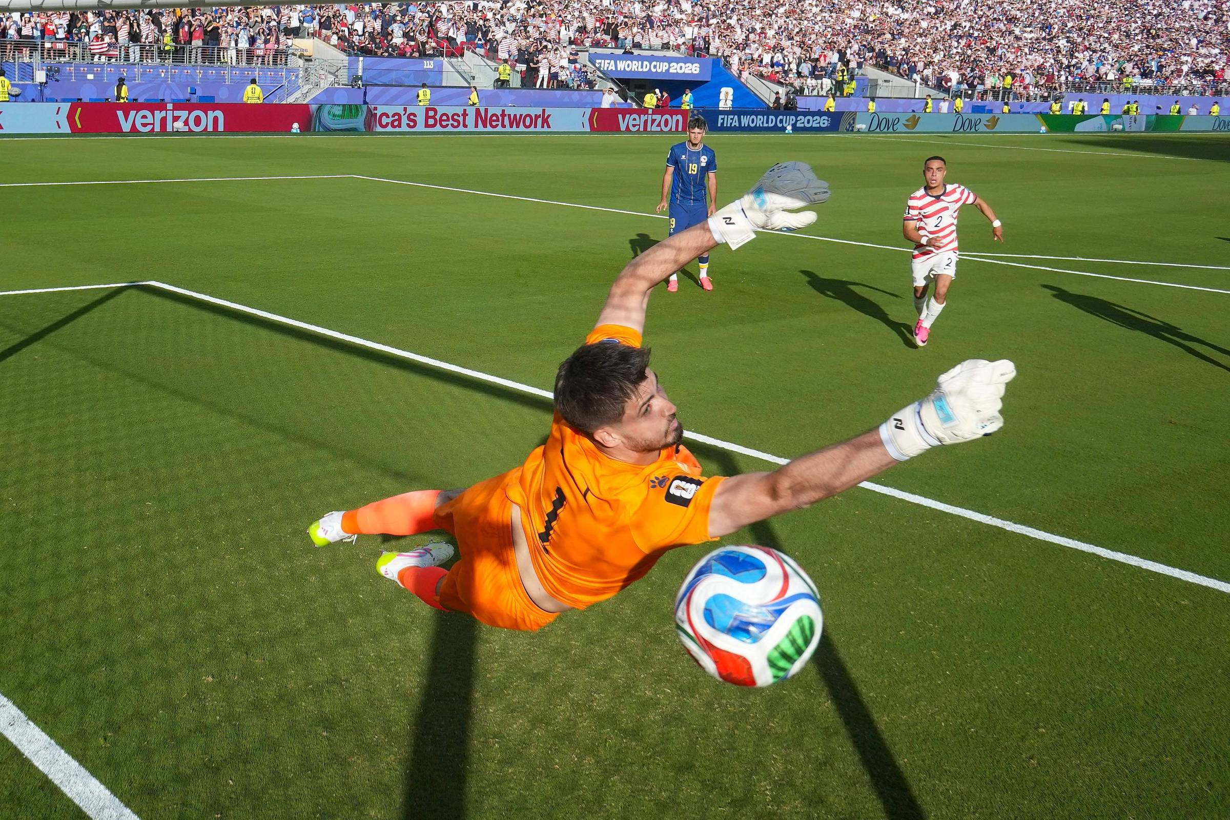 Goleiro vestido de laranja mergulha para a direita tentando defender chute de jogador adversário com uniforme branco e vermelho. Bola colorida está próxima às mãos do goleiro dentro da área, com torcida e outros jogadores ao fundo.