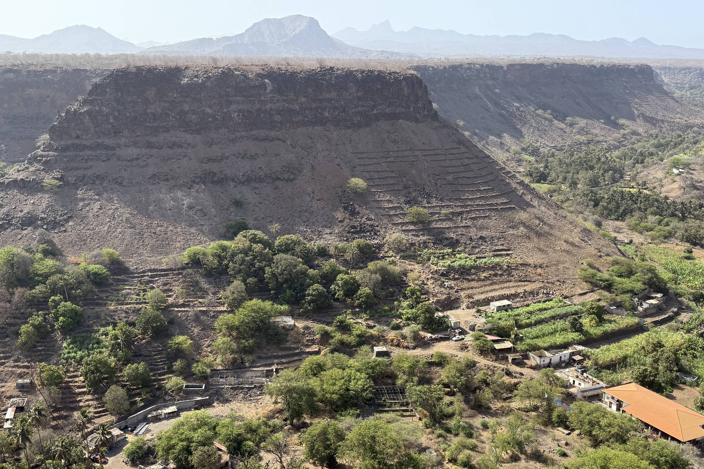 Vale com vegetação densa e algumas construções dispersas, cercado por morros rochosos de topo plano. Montanhas ao fundo sob céu claro.
