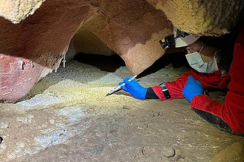 Pesquisador com máscara, luvas azuis e roupa vermelha usa lanterna e ferramenta para analisar inscrições em superfície rochosa dentro de caverna.