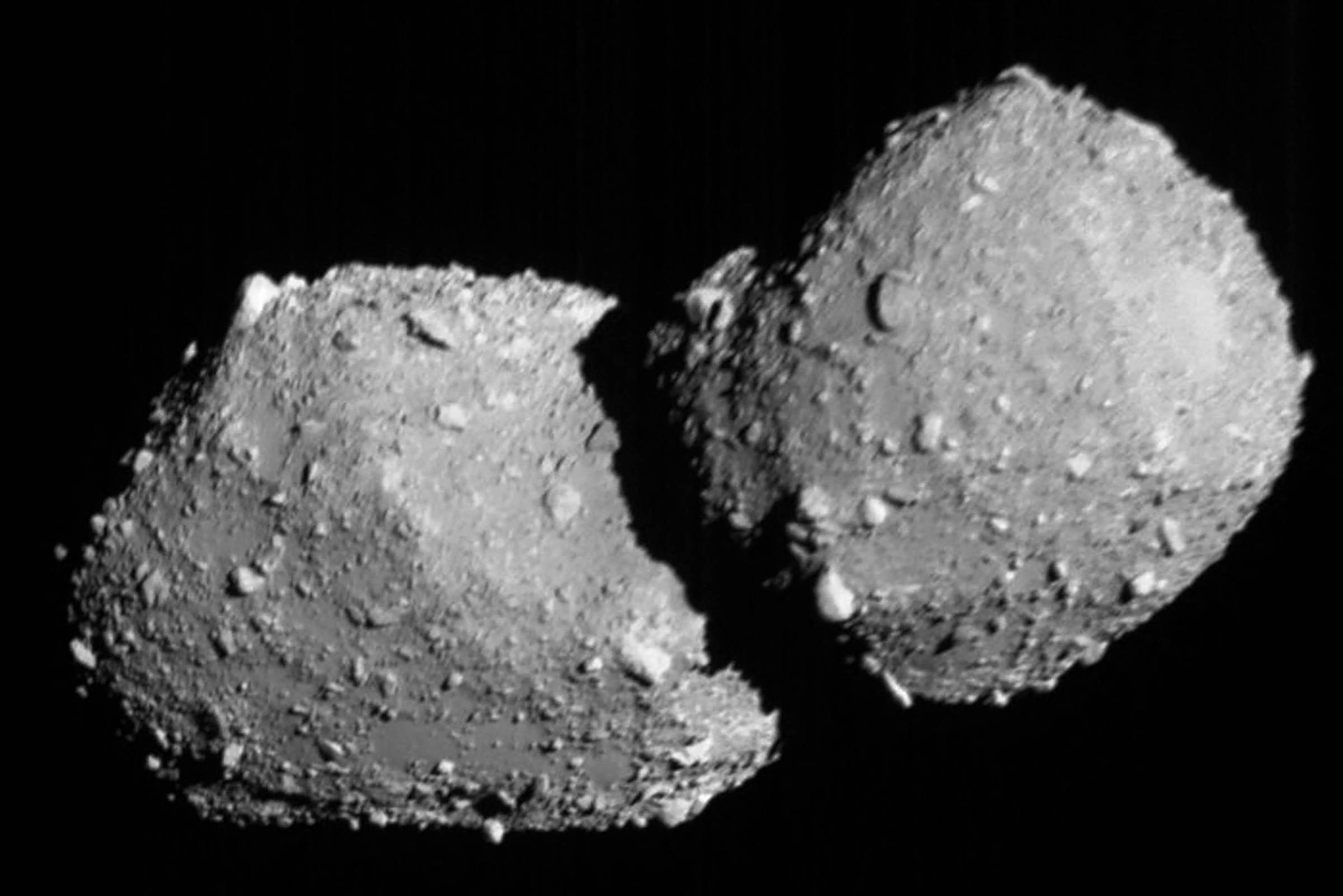 Asteroide binário composto por dois corpos rochosos irregulares conectados, com superfície cheia de crateras e fragmentos, em fundo preto do espaço.