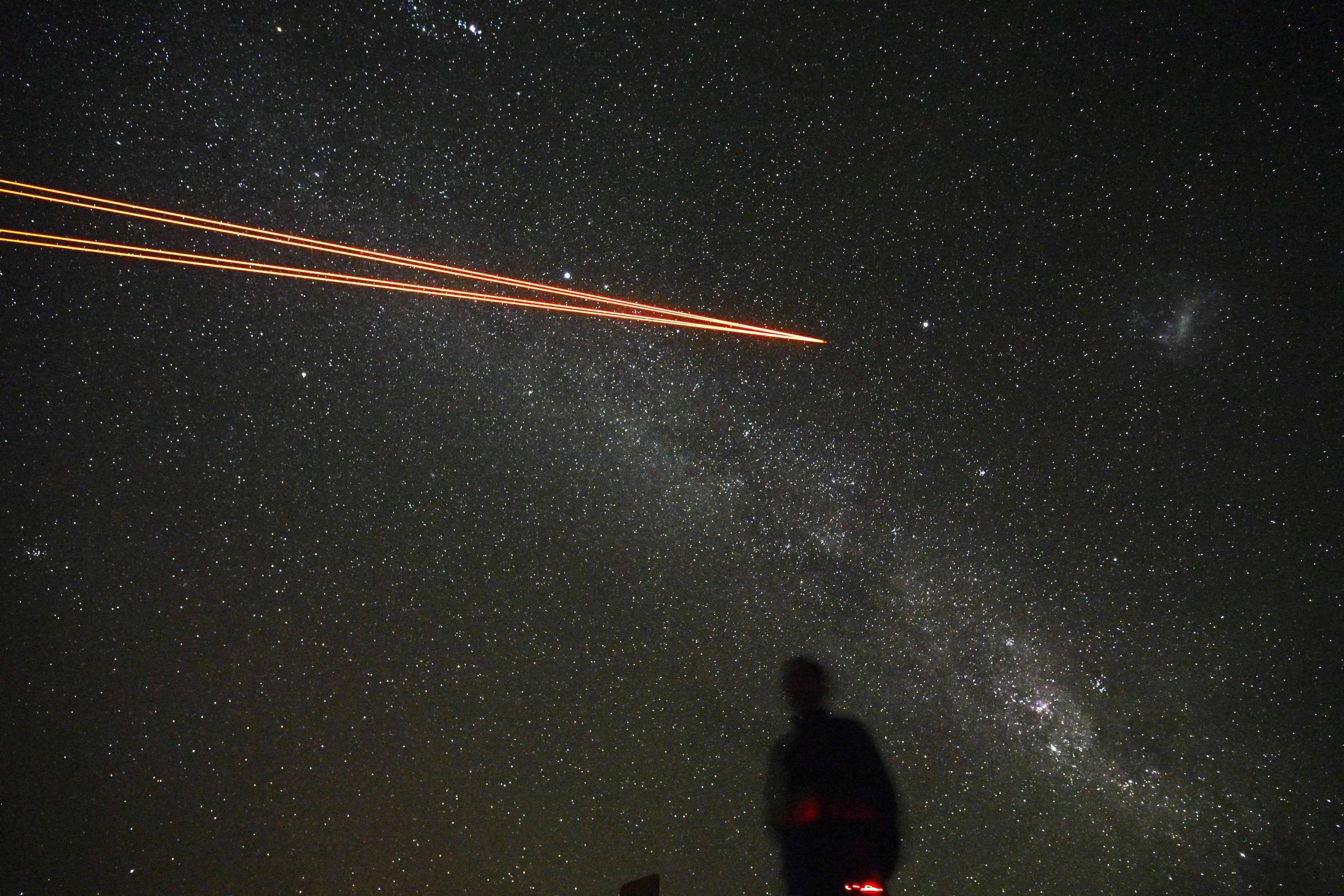 Céu noturno estrelado com a Via Láctea visível ao fundo. Dois rastros de luz laranja cruzam o céu da esquerda para a direita. No primeiro plano, silhueta escura de uma pessoa olhando para cima.