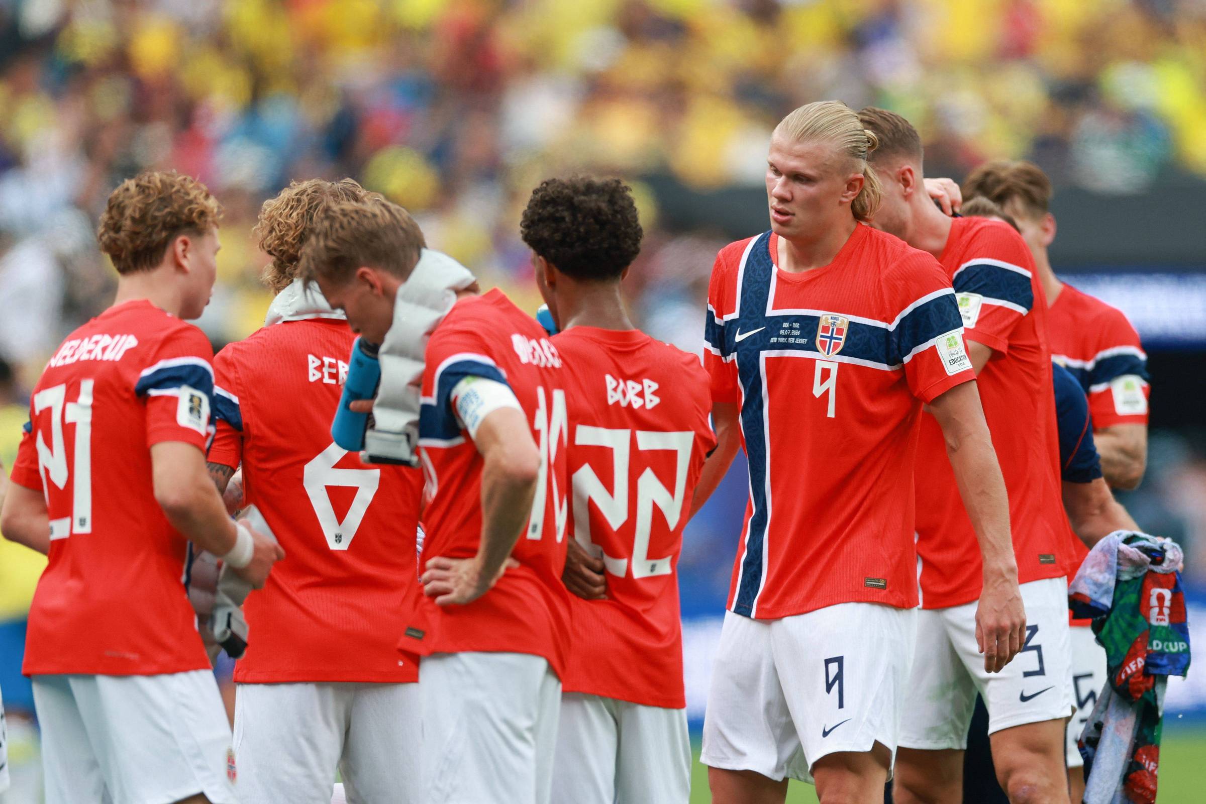 Seis jogadores da seleção norueguesa de futebol reunidos no campo durante intervalo. Eles vestem uniformes vermelhos com detalhes em azul e branco, e shorts brancos. Um jogador de cabelo loiro comprido destaca-se no centro da imagem.
