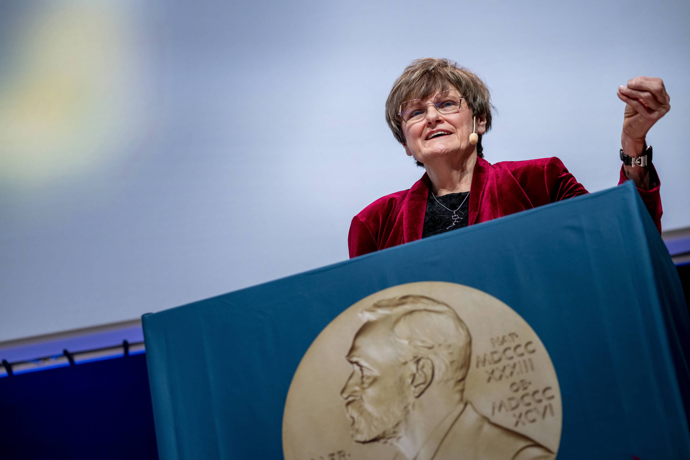 Mulher de cabelos curtos e óculos fala em púlpito coberto por tecido azul com medalha do Prêmio Nobel em relevo. Fundo neutro e iluminação suave.