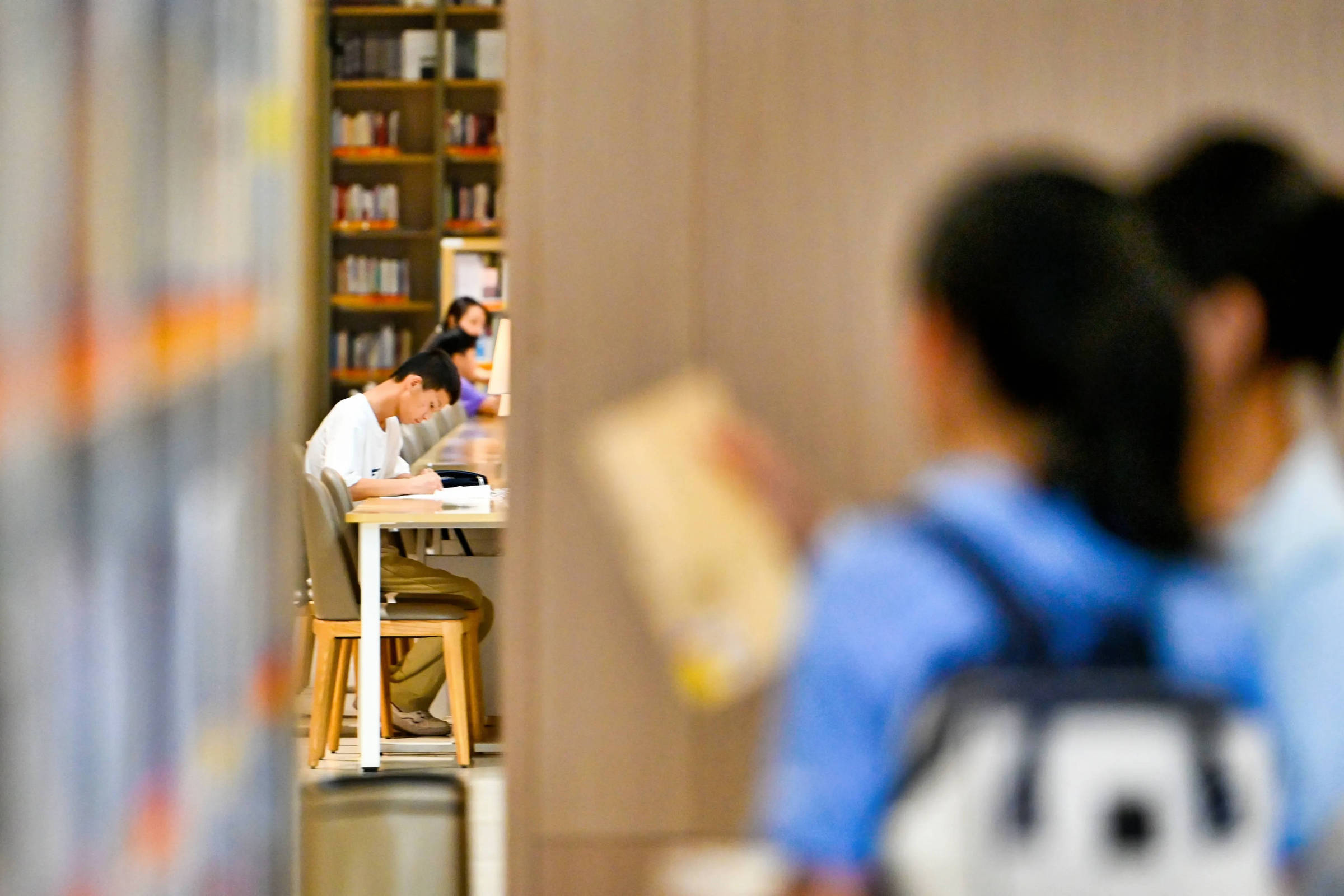 Dois estudantes sentados em mesa branca em corredor de biblioteca, focados na leitura. Prateleiras com livros ao fundo e outros estudantes em primeiro plano desfocados.