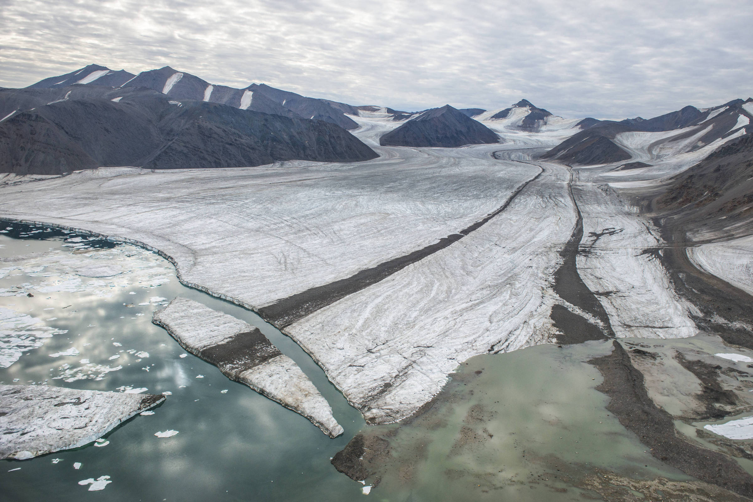 Glaciar extenso entre montanhas rochosas com áreas de gelo derretido formando lagos e canais de água. Céu nublado cobre a paisagem.