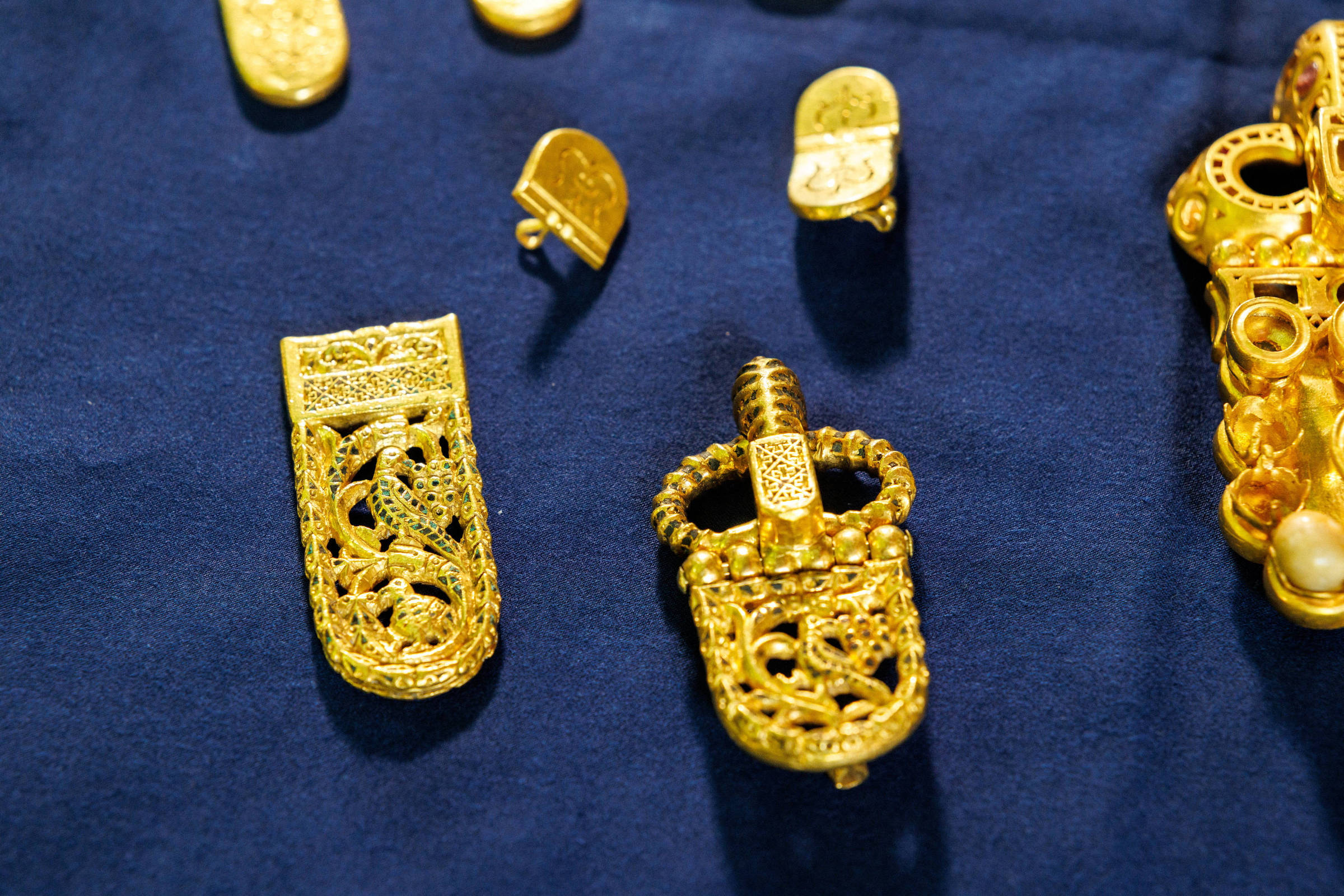 Peças de joias douradas com detalhes ornamentais complexos dispostas sobre tecido azul. Entre elas, há dois objetos maiores com desenhos vazados e texturas variadas, além de pequenos acessórios dourados ao redor.