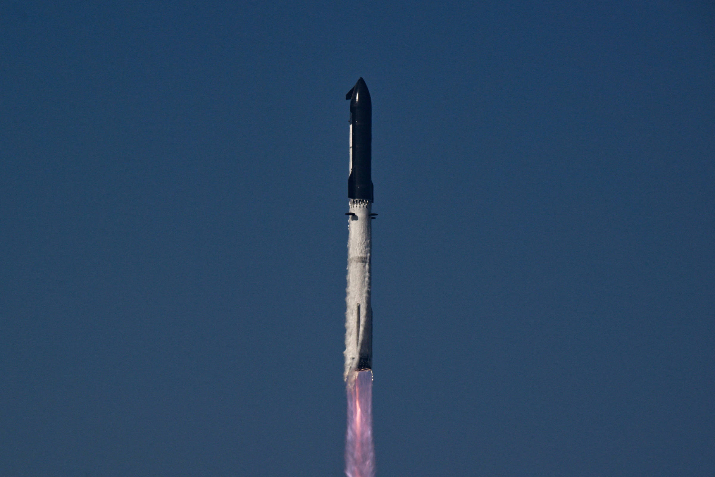 Foguete preto e branco em ascensão vertical com chamas rosadas saindo da base, contra céu azul sem nuvens.