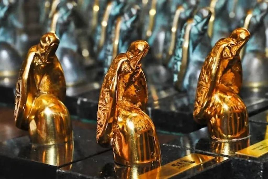 Três troféus dourados em forma de figura humana ajoelhada com capa, alinhados em base preta. Ao fundo, troféus semelhantes em tom prateado desfocados.