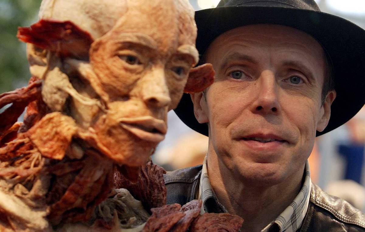 Homem de meia-idade usando chapéu preto e jaqueta de couro está ao lado de um modelo anatômico detalhado que mostra músculos e tecidos humanos sem pele.