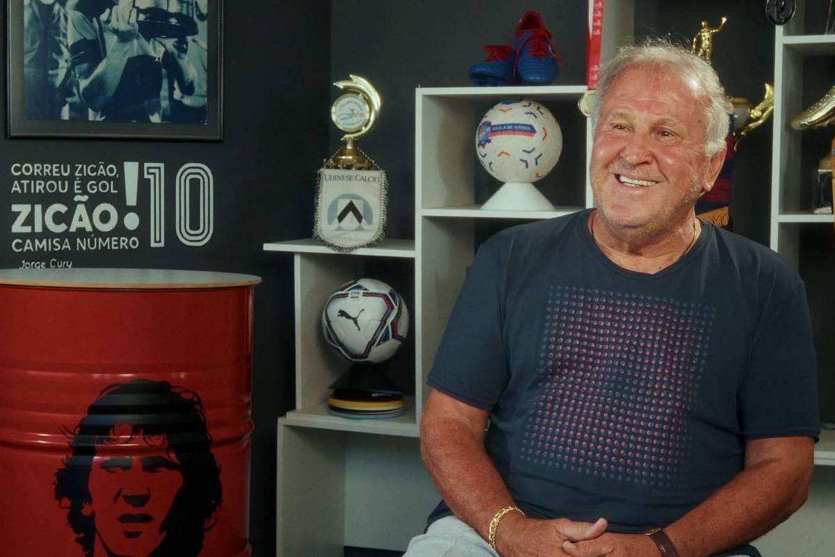 Homem idoso de cabelo branco e camiseta azul escura sorri sentado em sala com prateleiras ao fundo contendo troféus, bolas de futebol e medalhas. À esquerda, tambor vermelho com imagem estilizada de rosto e texto sobre Zico e número 10.