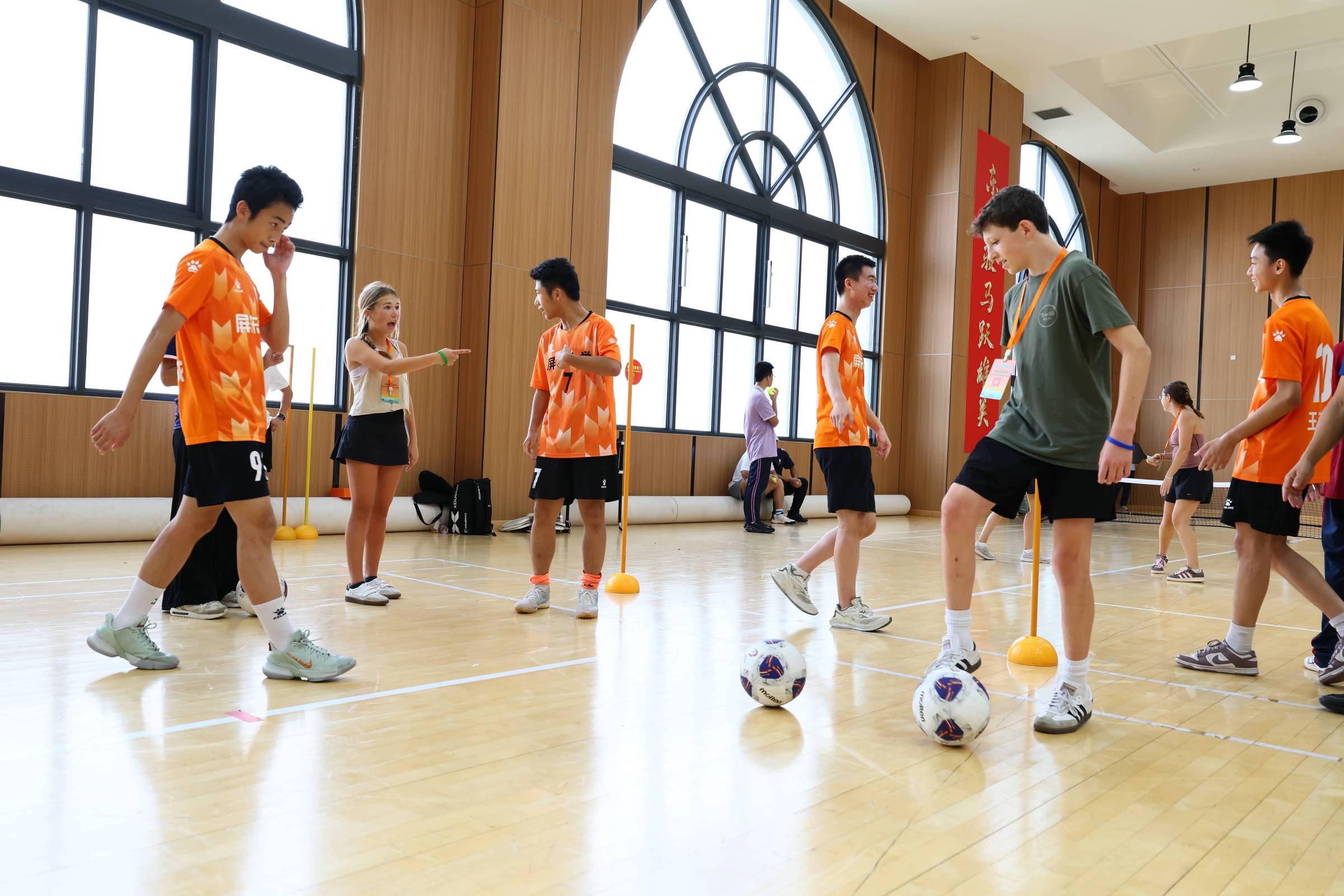 Seis jovens atletas com uniformes laranja e shorts pretos treinam em quadra coberta com piso de madeira. Dois deles controlam bolas de futebol próximas a cones laranja, enquanto uma mulher orienta e aponta para um dos jogadores. Grandes janelas arqueadas iluminam o ambiente interno.