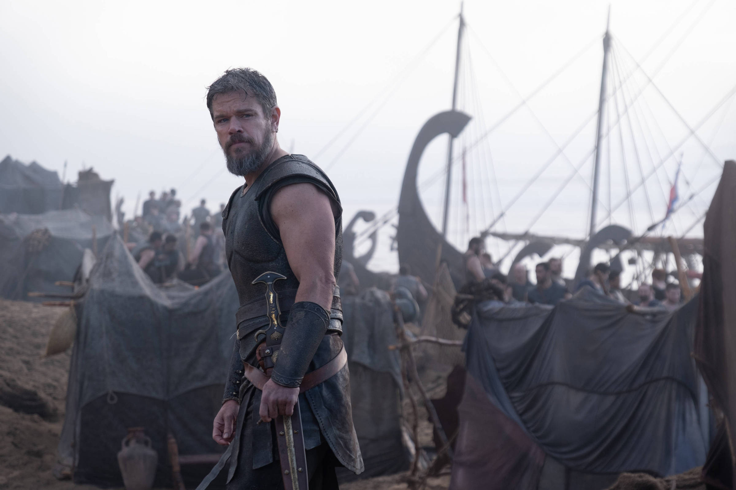 Homem vestido com armadura de couro preta e espada na cintura está em primeiro plano, olhando para a câmera. Ao fundo, grupo de pessoas e um navio viking com velas e mastros visíveis em ambiente enevoado.
