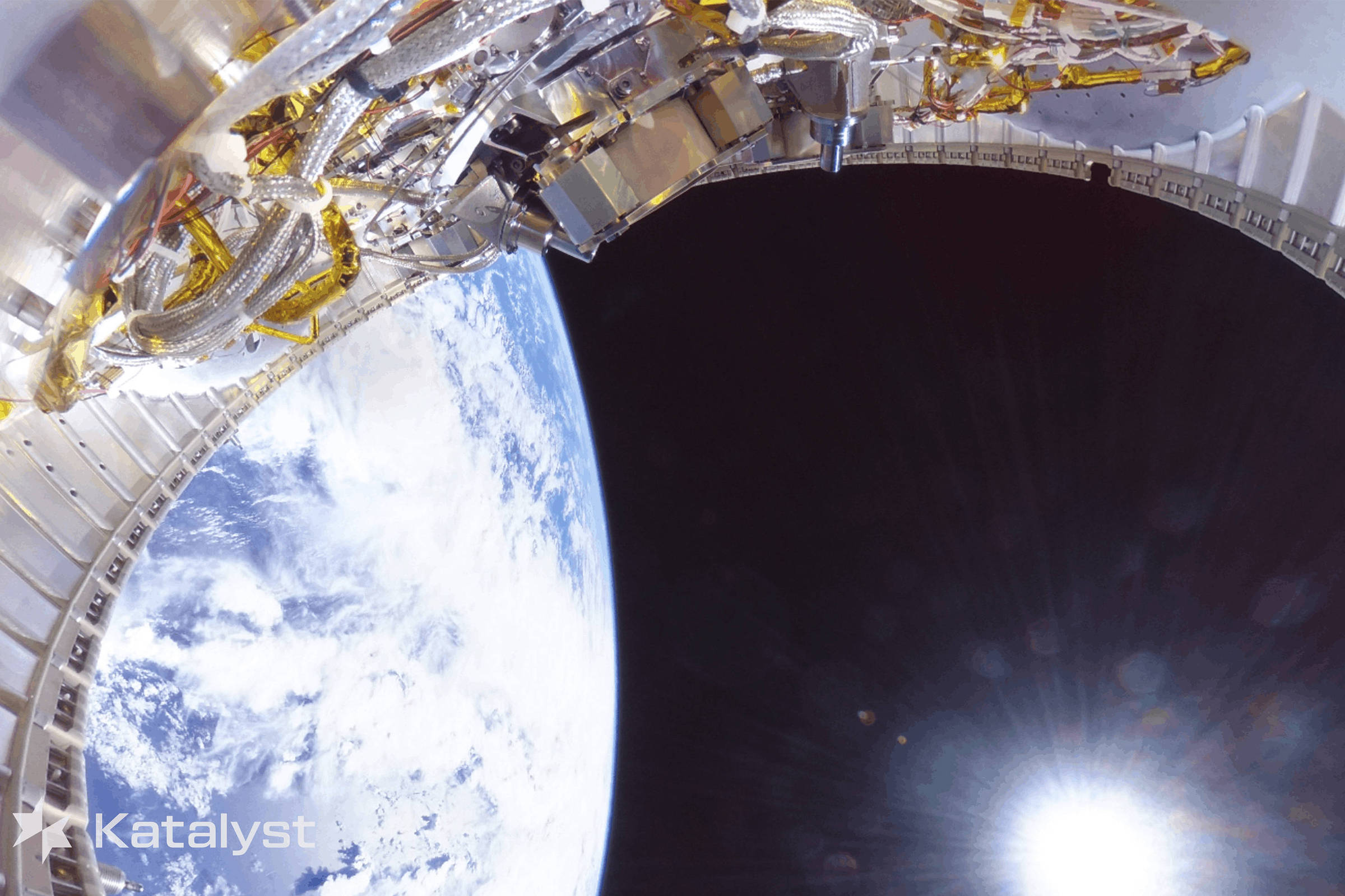 Parte de um módulo espacial com componentes metálicos e cabos visível no canto superior esquerdo. Abaixo, a Terra aparece com nuvens brancas sobre o oceano azul. O espaço escuro e o sol brilhante estão à direita.