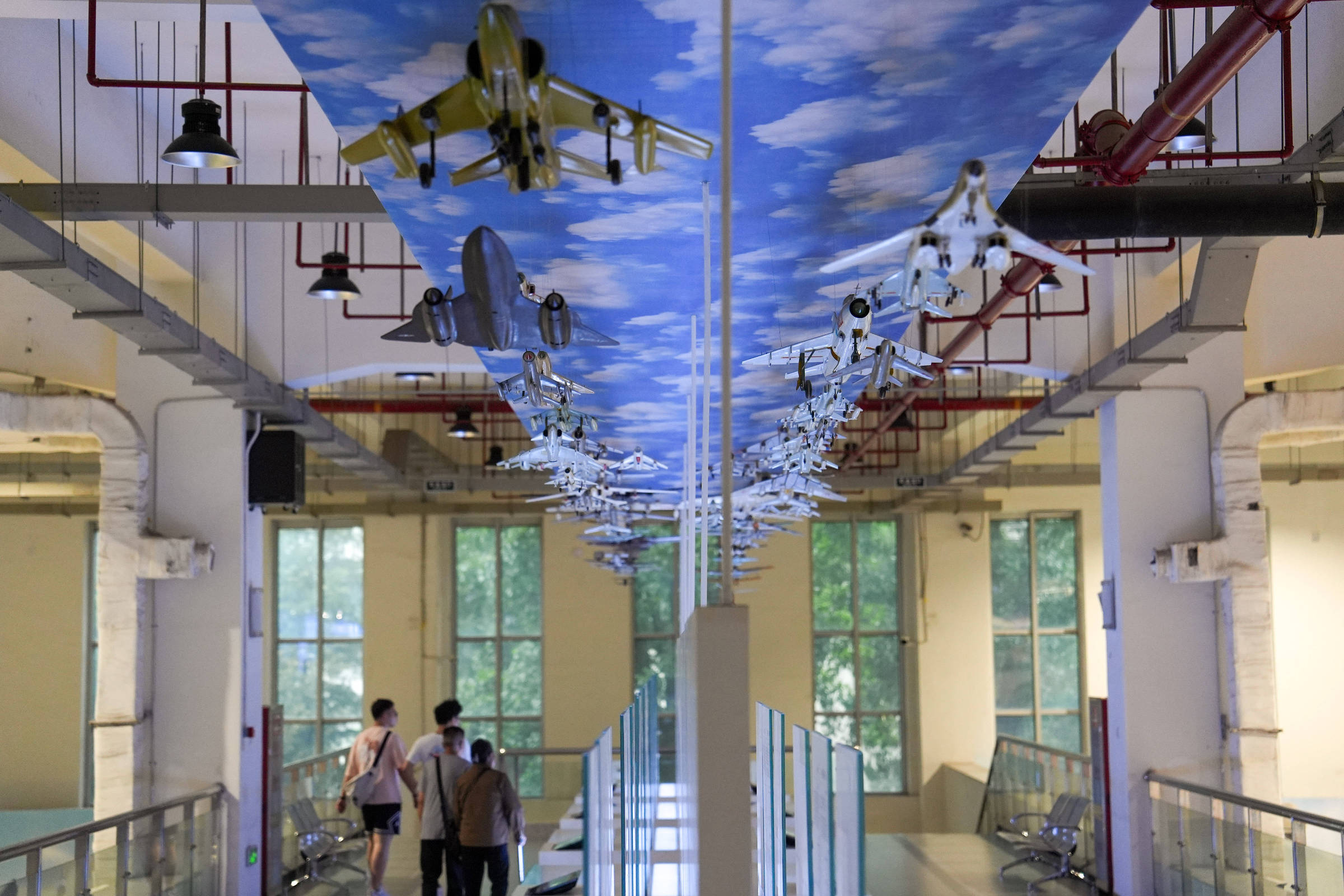 Corredor interno de museu com teto decorado por painéis azuis com nuvens e modelos de aviões suspensos em fileira central. Três visitantes caminham pelo corredor entre paredes claras e janelas grandes ao fundo.