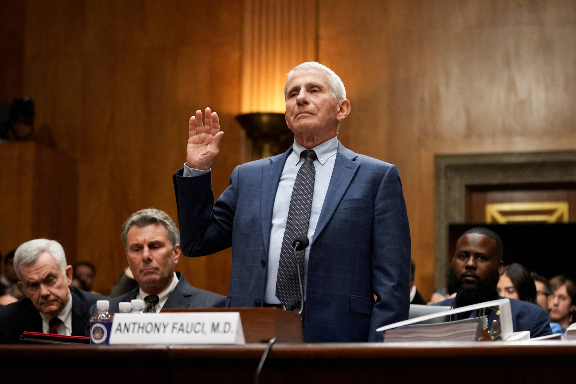Anthony Fauci em pé, com a mão direita levantada, prestando juramento em audiência pública. Ele veste terno azul e gravata, com microfone à sua frente e placa com seu nome. Pessoas sentadas ao fundo observam a cena em ambiente formal de madeira.