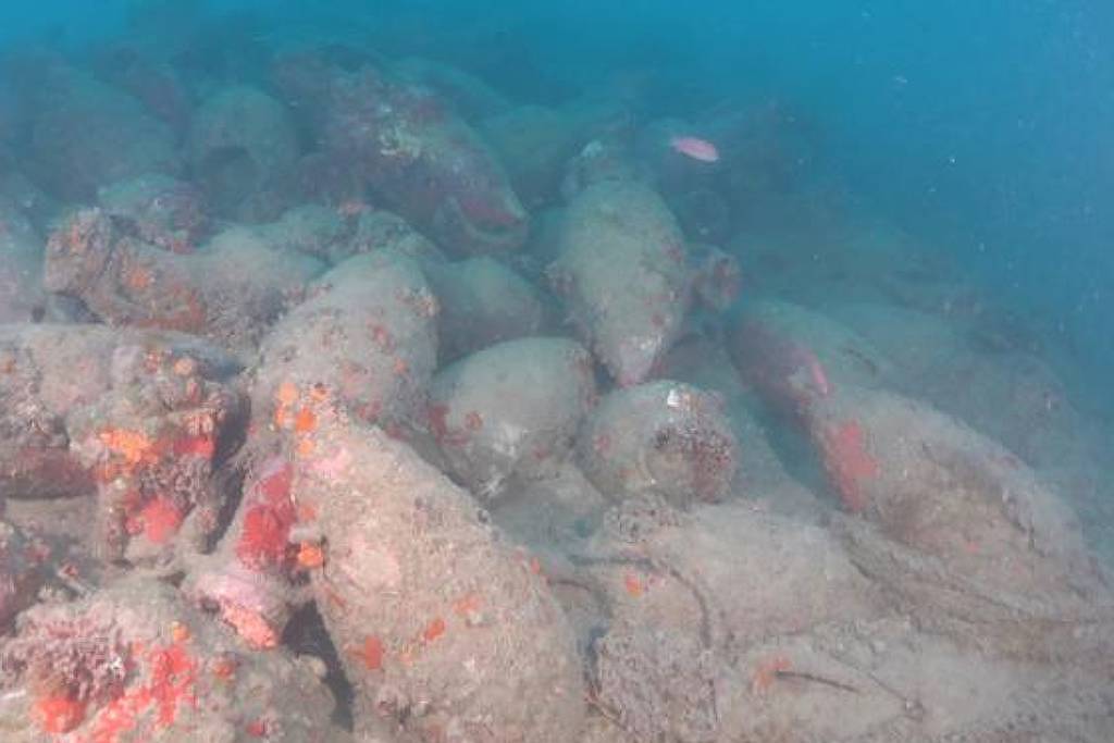 Diversas ânforas antigas cobertas por sedimentos e organismos marinhos repousam no fundo do mar, parcialmente enterradas em areia e visíveis em água azulada.