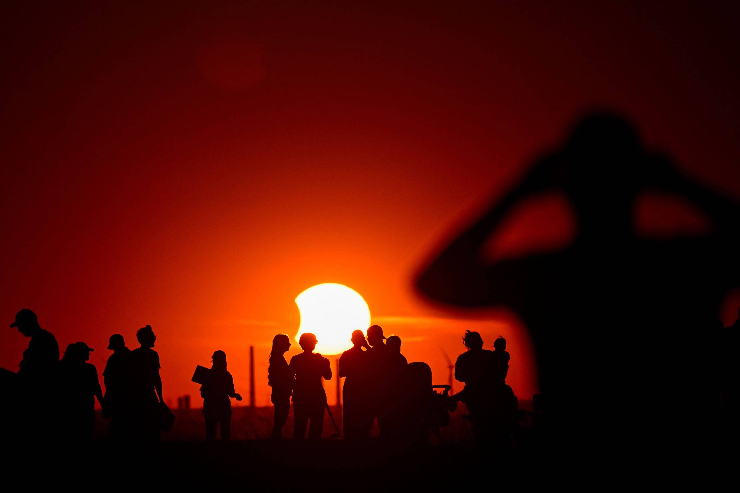 Silhuetas de várias pessoas observam o eclipse solar parcial no horizonte durante o pôr do sol, com o céu em tons intensos de vermelho e laranja.