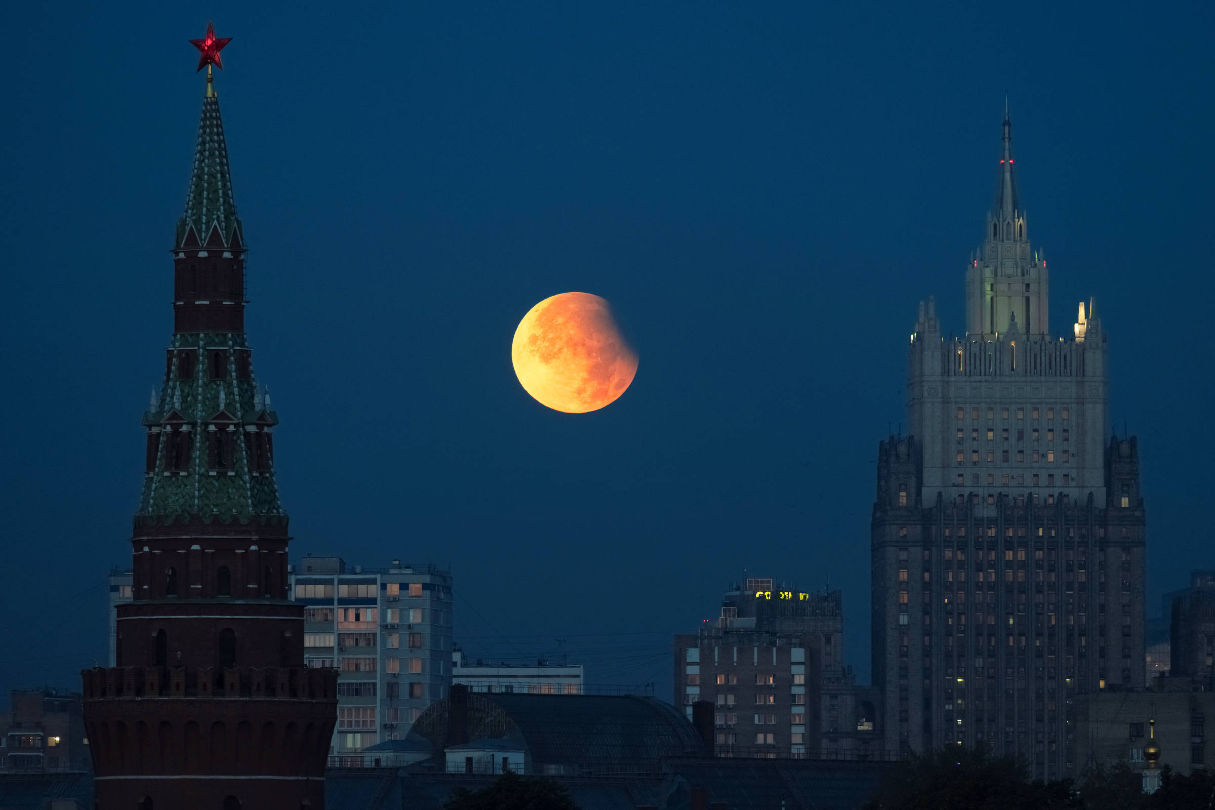 Lua parcialmente eclipsada aparece no céu noturno entre duas torres icônicas da cidade de Moscou, com iluminação artificial nas construções ao redor.