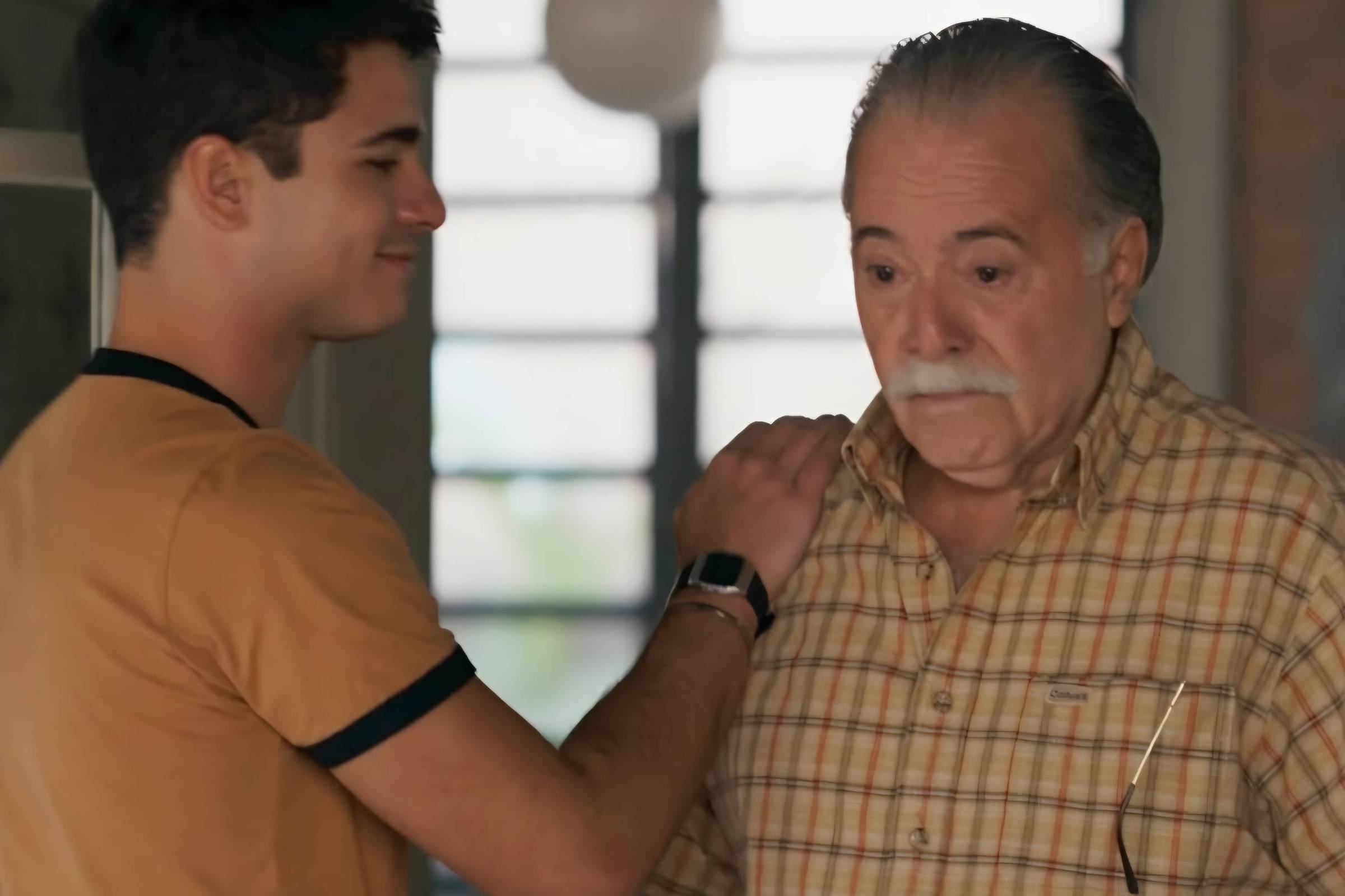 Homem jovem com camiseta marrom clara toca o ombro de homem idoso de camisa xadrez bege, ambos em ambiente interno com janela ao fundo.