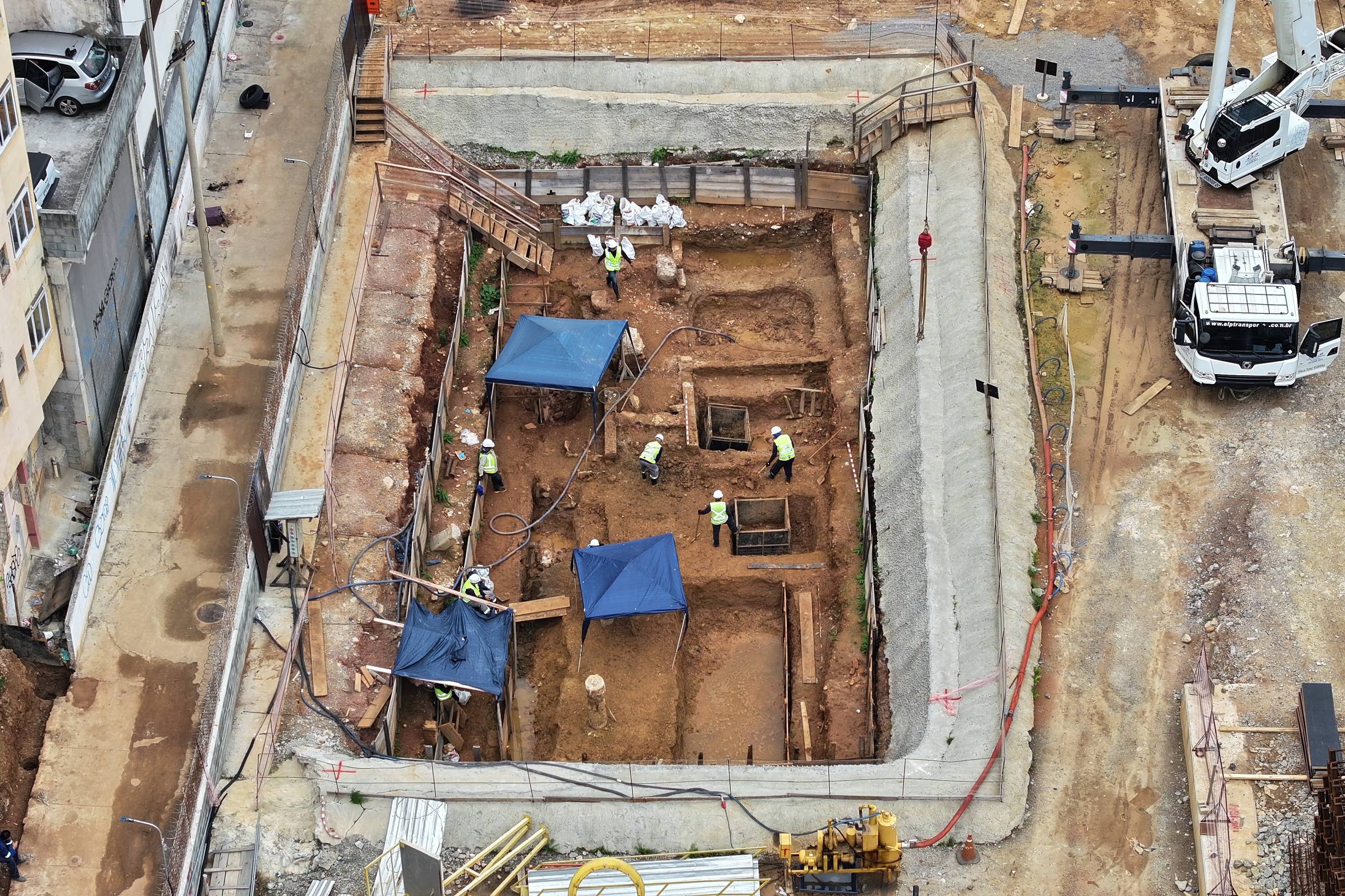 Vista aérea mostra canteiro de obras com escavações retangulares cercadas por tapumes. Cinco trabalhadores com coletes refletores estão no local, com duas tendas azuis para sombra. Equipamentos de construção, incluindo guindaste, estão posicionados ao redor da área.