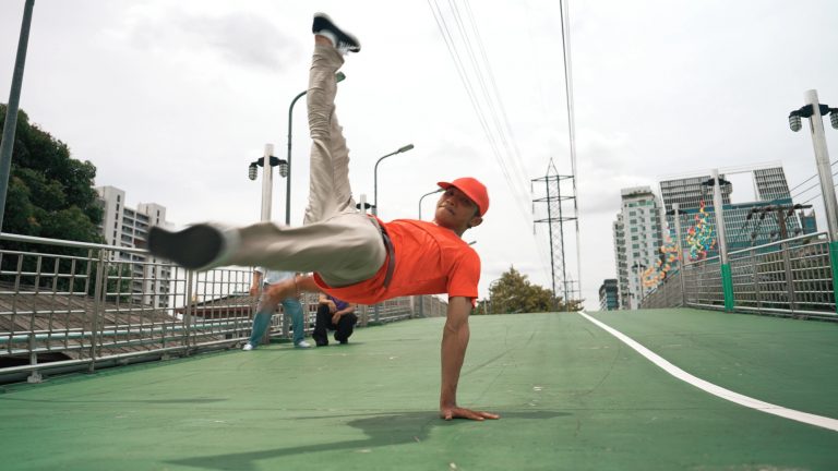 Um dançarino de boné e camiseta vermelhos executa um movimento de breakdance, apoiado em uma das mãos e com as pernas erguidas. A apresentação ocorre em uma passarela urbana de piso verde, cercada por grades, postes, prédios e fios de alta tensão, sob o céu nublado