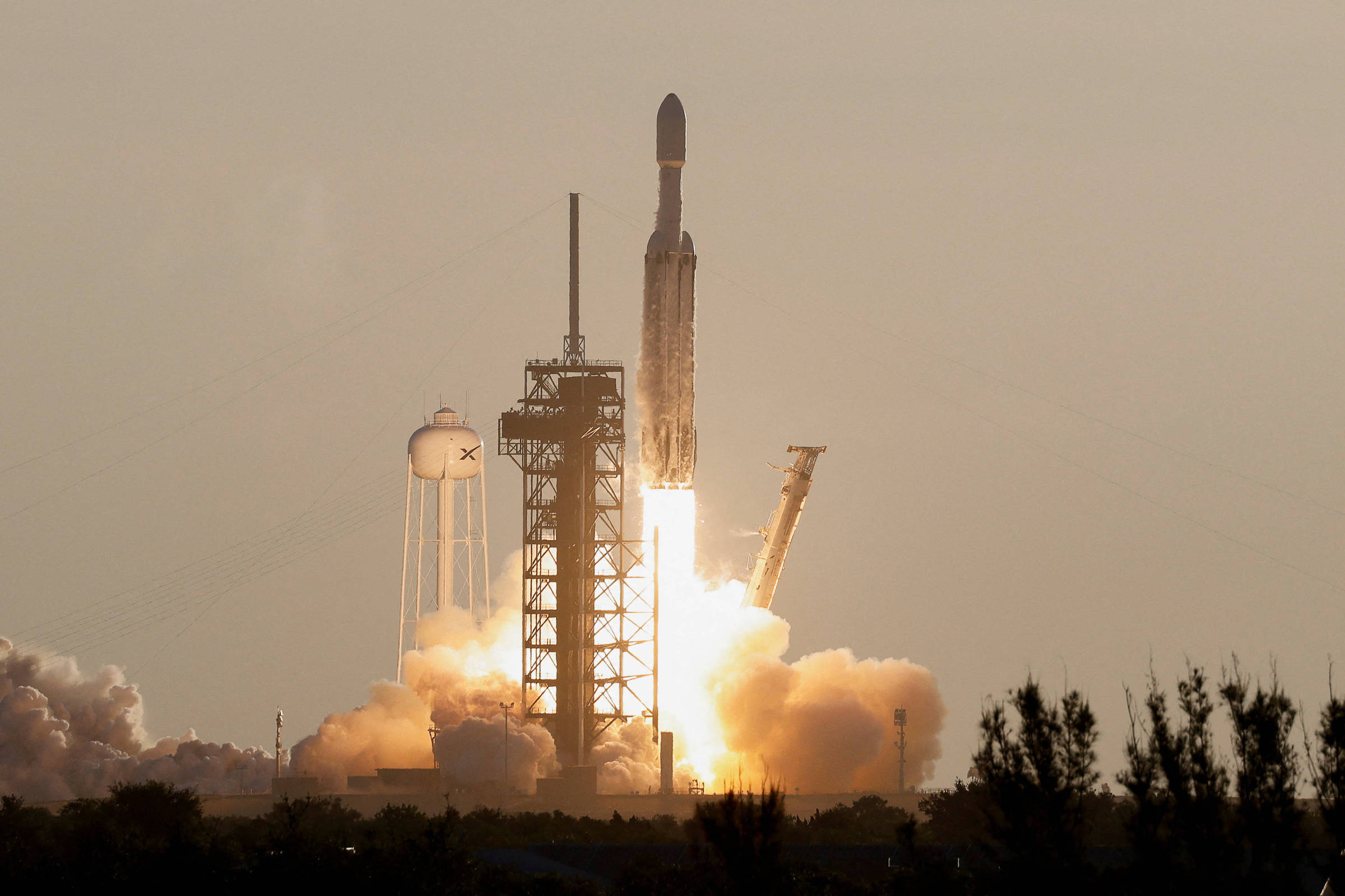 Foguete Falcon Heavy decola de plataforma de lançamento com chamas e fumaça intensa ao entardecer. Estruturas da base e vegetação aparecem em primeiro plano.