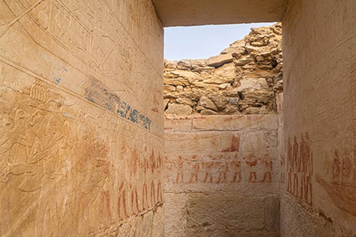 Corredor estreito em ruínas arqueológicas com paredes de pedra decoradas por relevos e pinturas antigas em tons avermelhados e ocres. No topo, céu azul visível acima de pedras empilhadas.