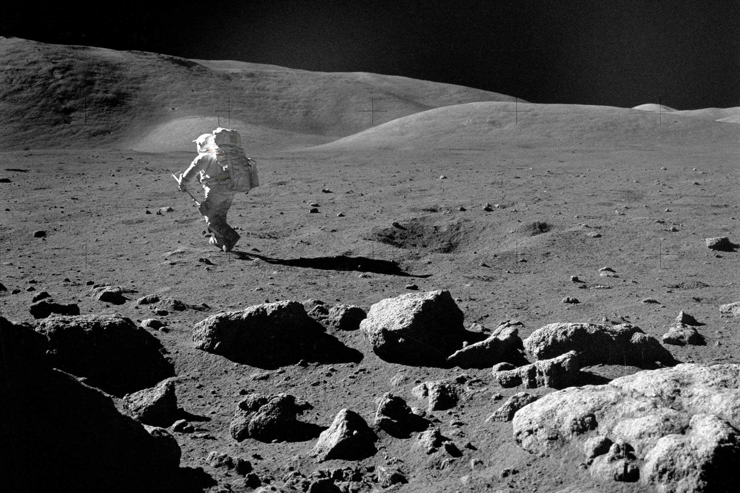 Astronauta em traje espacial completo caminha sobre terreno rochoso e irregular da Lua, com colinas suaves ao fundo e céu negro sem estrelas.