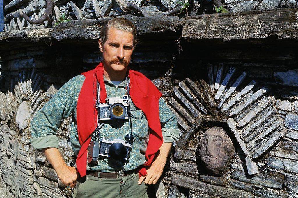 Homem com bigode e cabelo penteado usa duas câmeras fotográficas penduradas no pescoço, veste camisa verde e suéter vermelho sobre os ombros. Ele está em frente a uma parede de pedra com decoração em formato de arco e rosto esculpido.