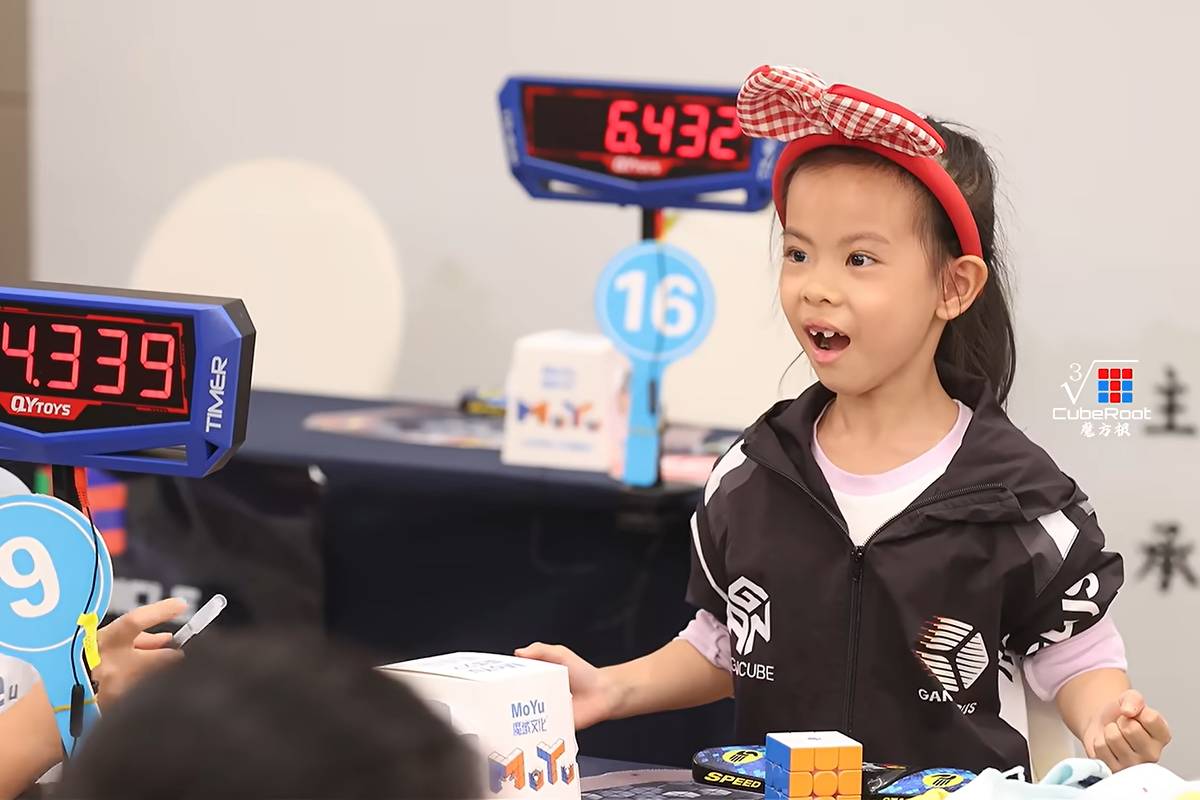 Menina com faixa vermelha na cabeça segura cubo mágico em competição. Placas digitais mostram tempos 4,339 e 6,432 segundos.