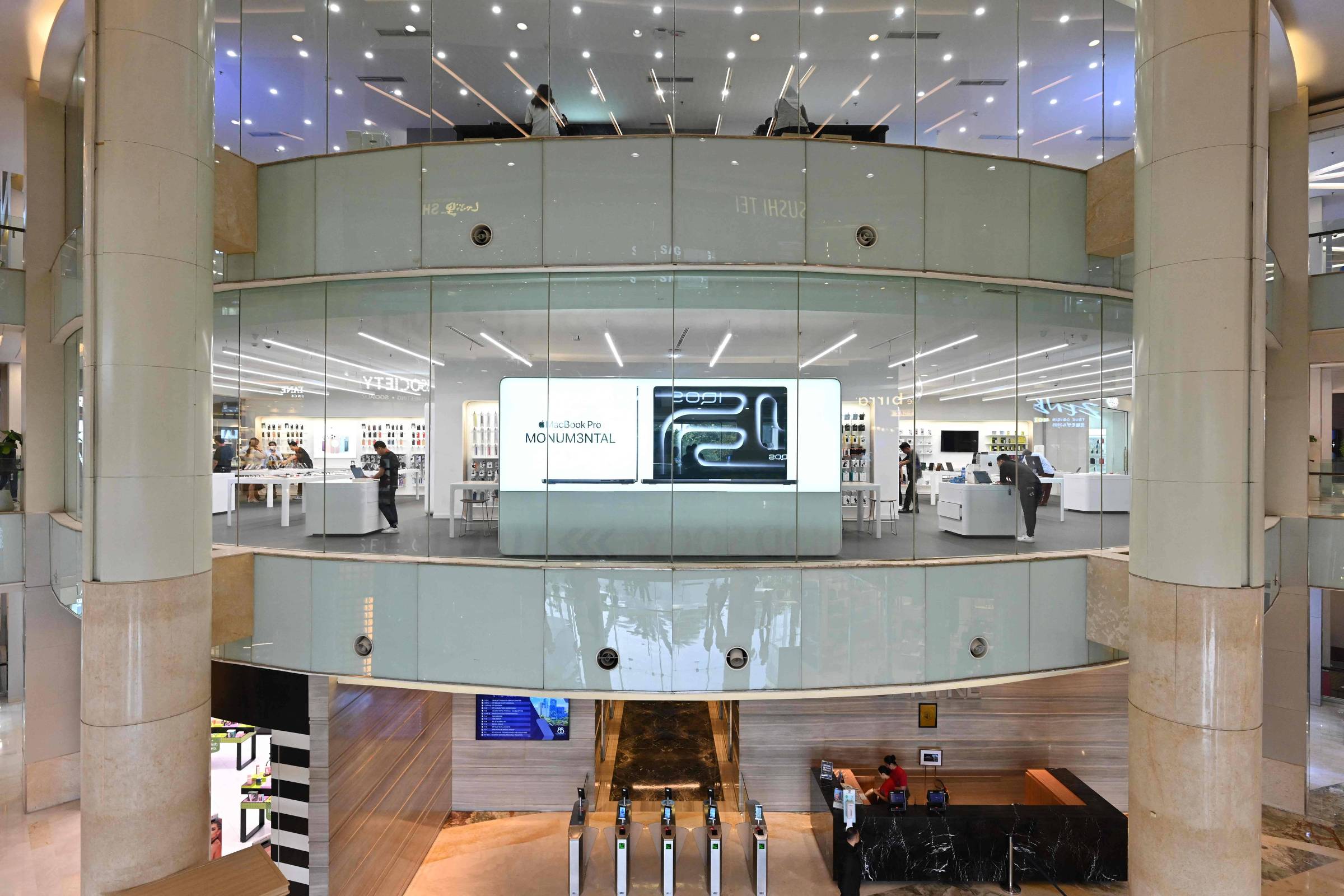 Loja Apple com fachada de vidro em dois andares dentro de shopping. No andar superior, painéis digitais exibem produtos e logotipo da Apple. Pessoas circulam e atendem clientes. Escada rolante conecta ao térreo, onde há balcão de atendimento e área de espera.