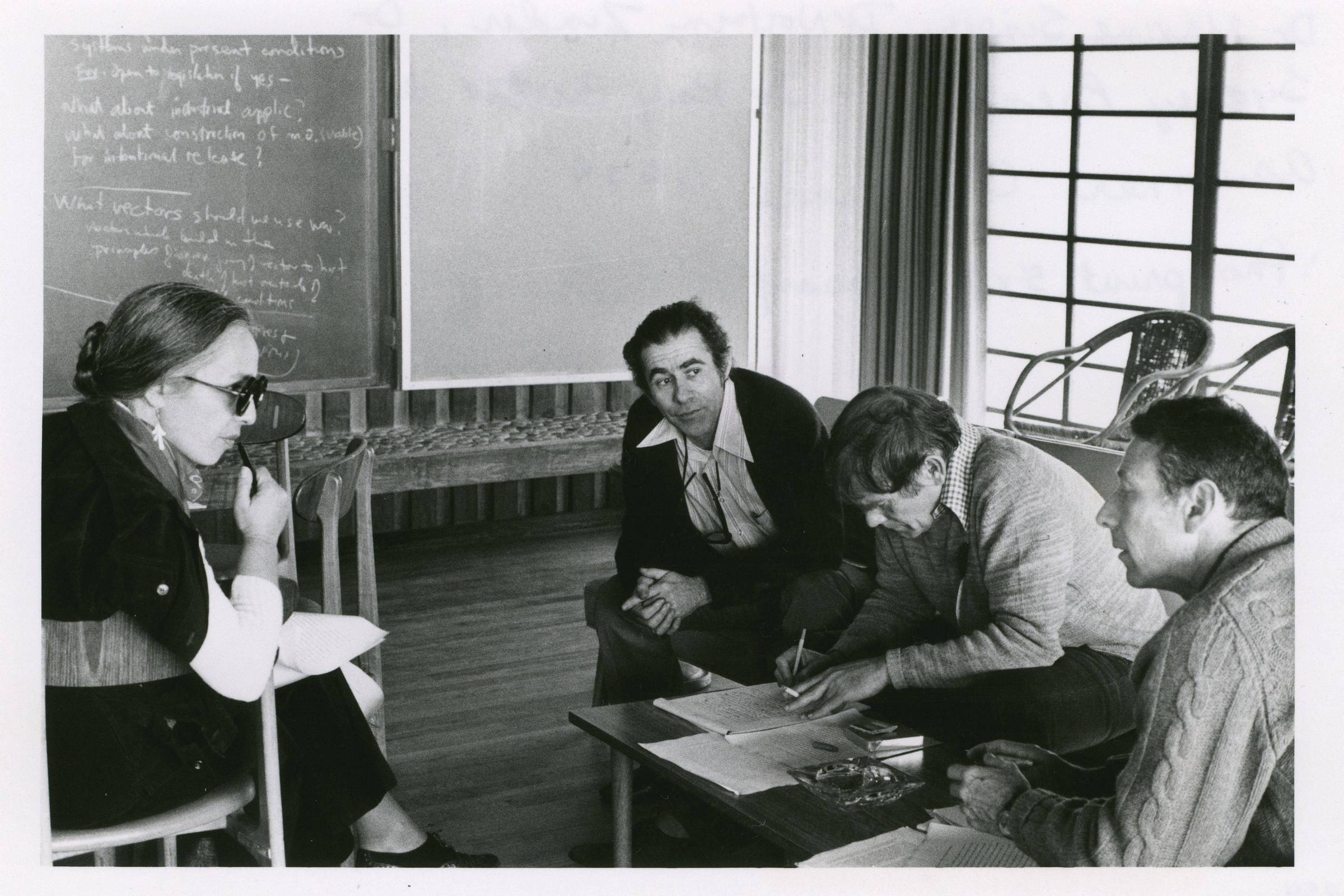 Quatro adultos sentados em sala discutem documentos sobre mesa baixa. Mulher à esquerda usa óculos escuros e segura papel. Três homens à direita observam e escrevem em papéis. Quadro-negro com anotações ao fundo e janela com cortinas à direita.