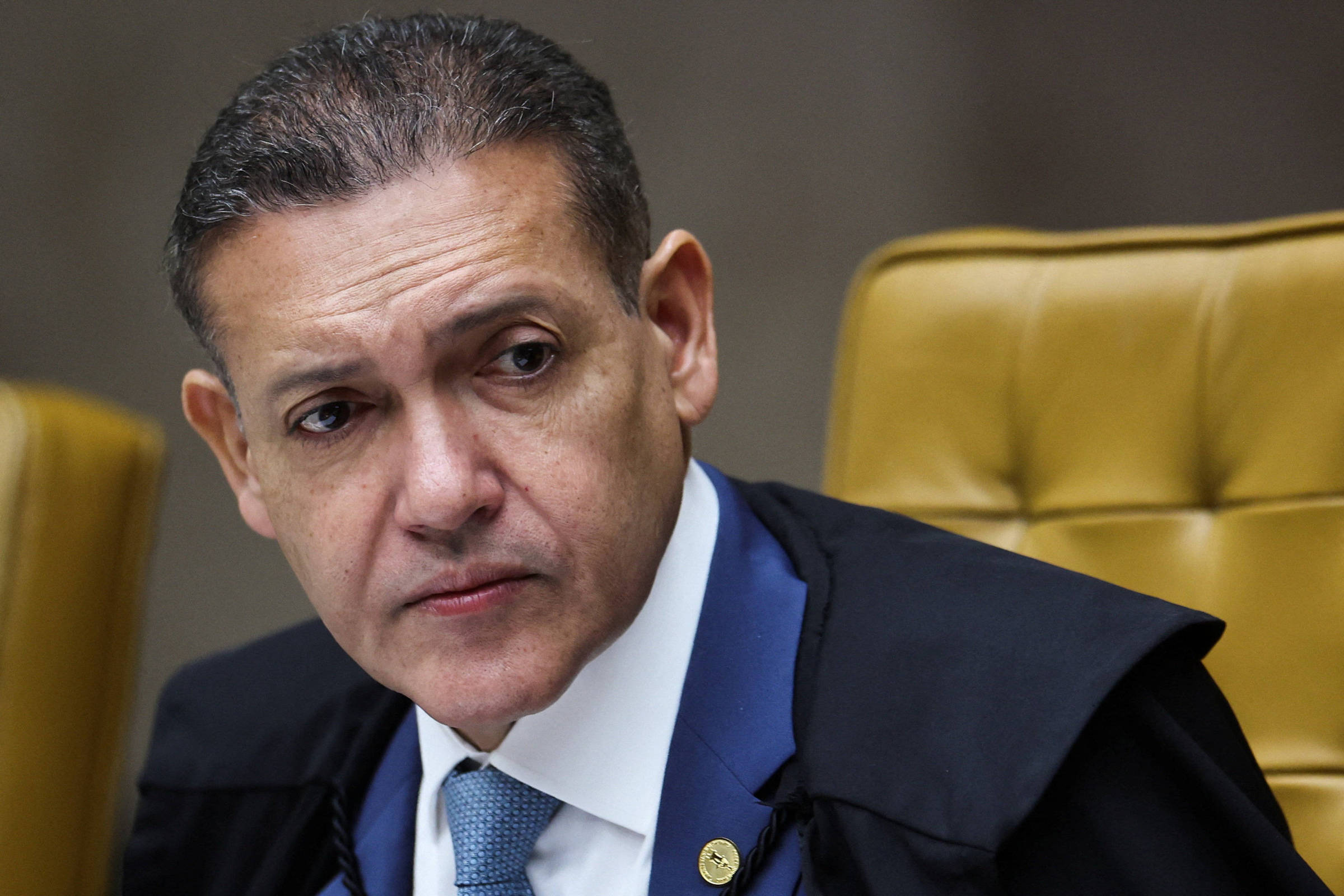 Homem de meia-idade com cabelo curto e escuro veste terno azul, camisa branca e gravata azul clara, sentado em cadeira amarela acolchoada, olhando para o lado.