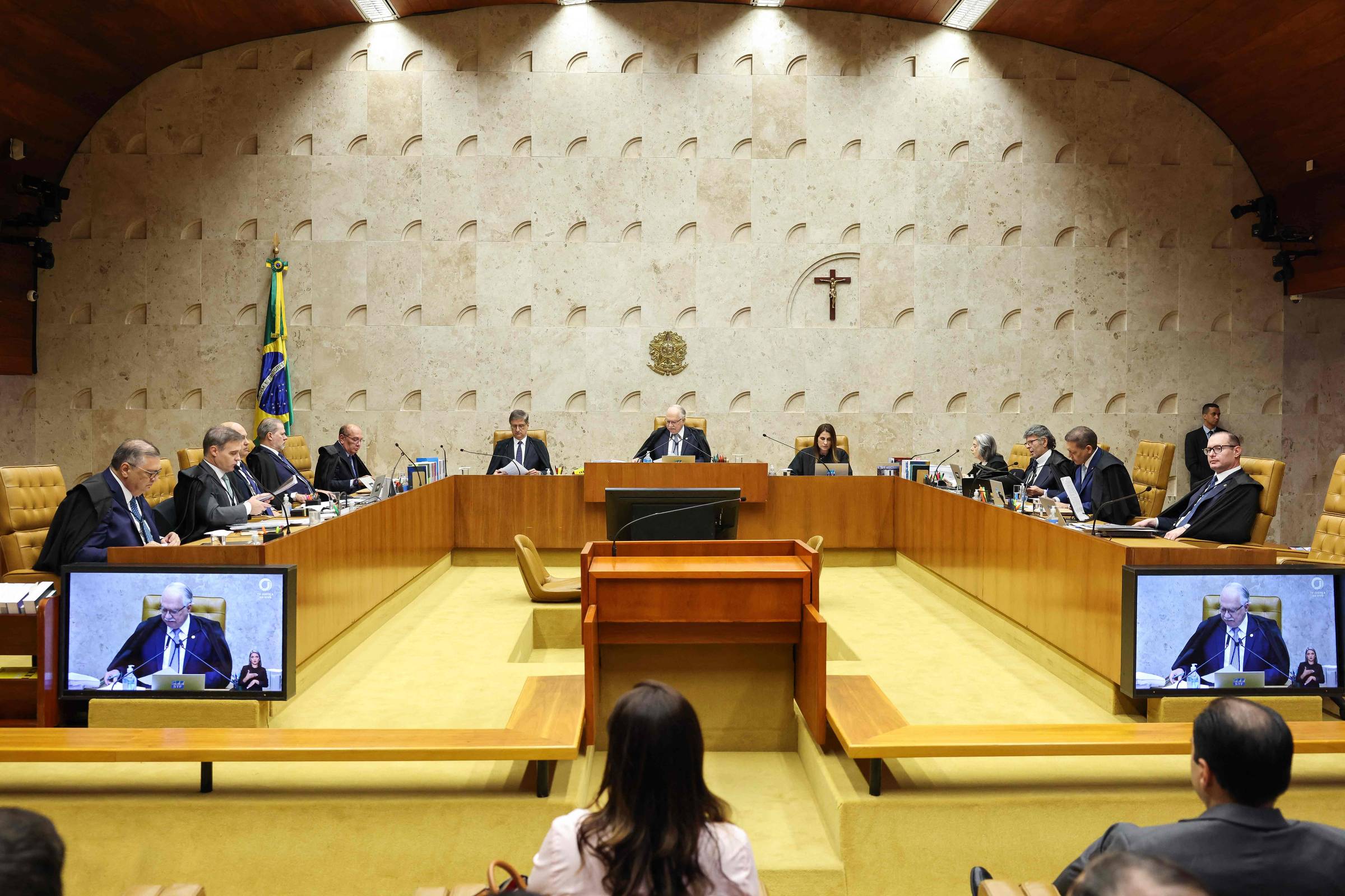 Ministros sentados em semicírculo no plenário do Supremo Tribunal Federal, com bandeira do Brasil e crucifixo na parede ao fundo. Duas telas exibem imagem de um dos ministros falando.