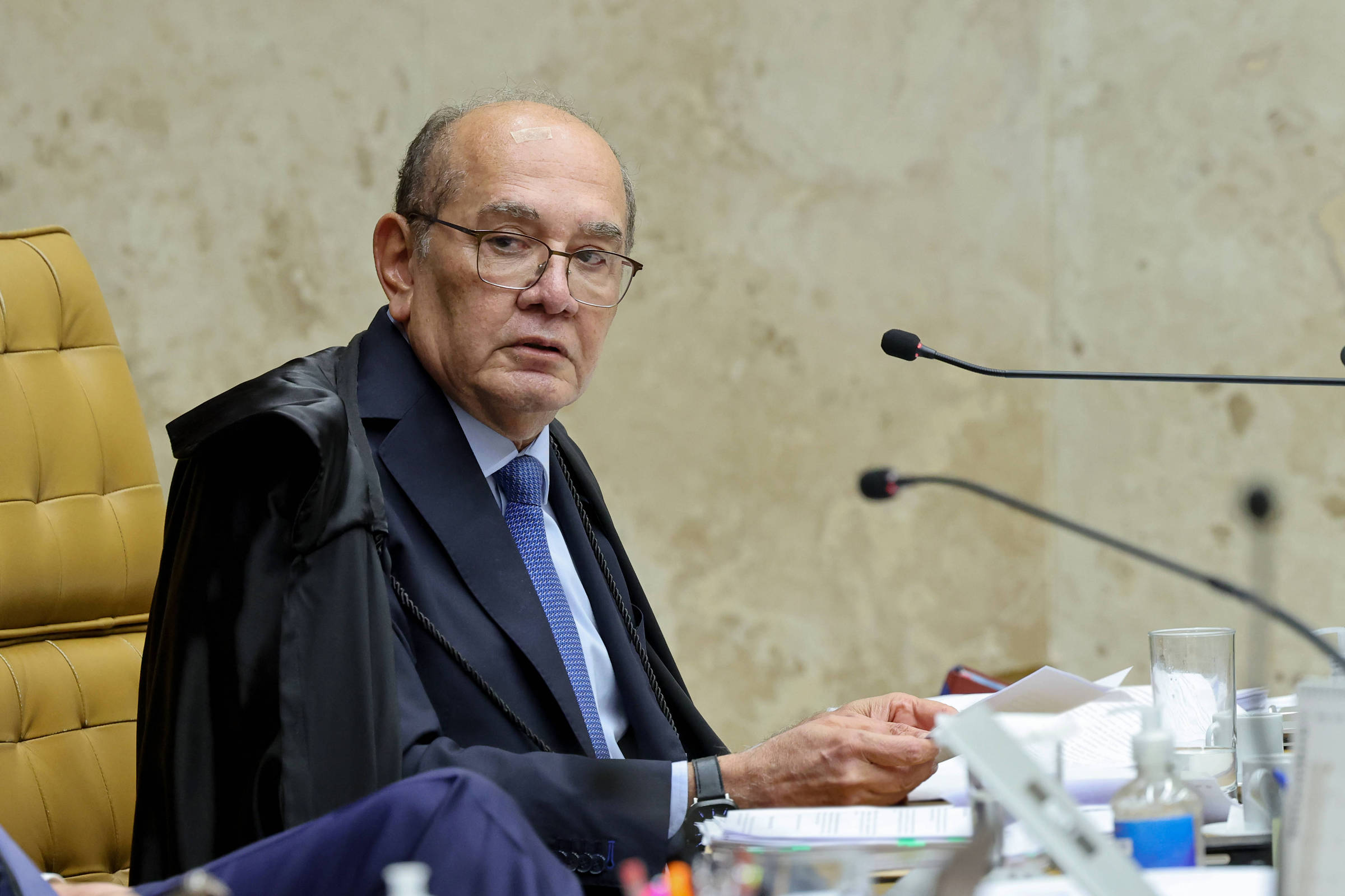 Senhor com óculos e terno azul escuro, usando toga preta, sentado em cadeira amarela em ambiente de tribunal. Ele segura papéis e está diante de microfones e objetos de escritório sobre a mesa.
