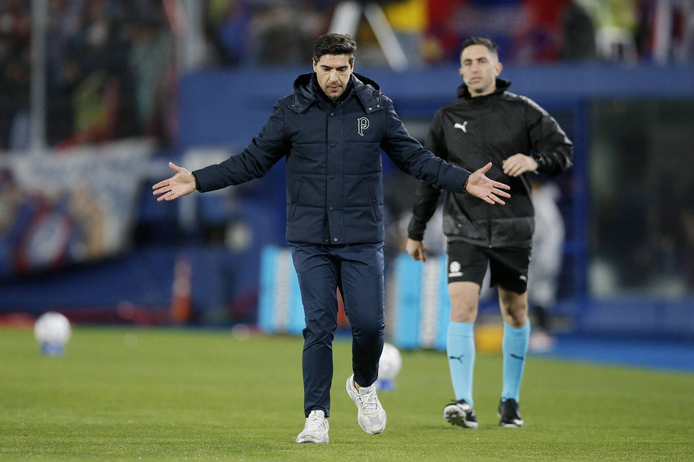 Treinador com casaco azul escuro e calça azul gesticula com os braços abertos no gramado. Árbitro vestido de preto e azul claro caminha atrás dele. Fundo desfocado com público e elementos do estádio.